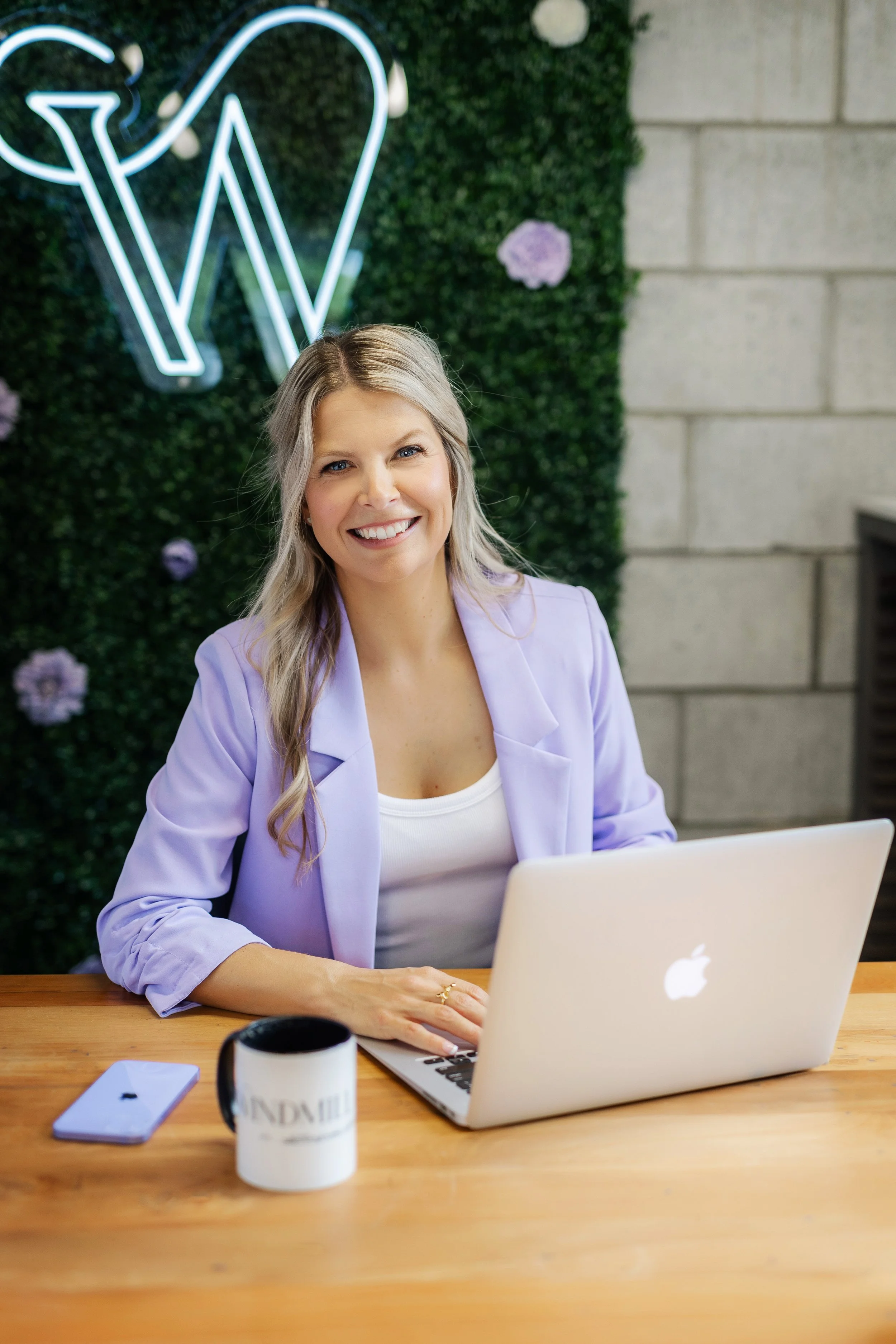 A woman with blonde hair, smiling, sitting at a wooden desk with a silver MacBook laptop open, a coffee mug, a smartphone, and a closed notebook. She is wearing a light purple blazer and a white top. Behind her, there is a green wall with purple flowers and a neon light sign, and a brick wall on the right side.