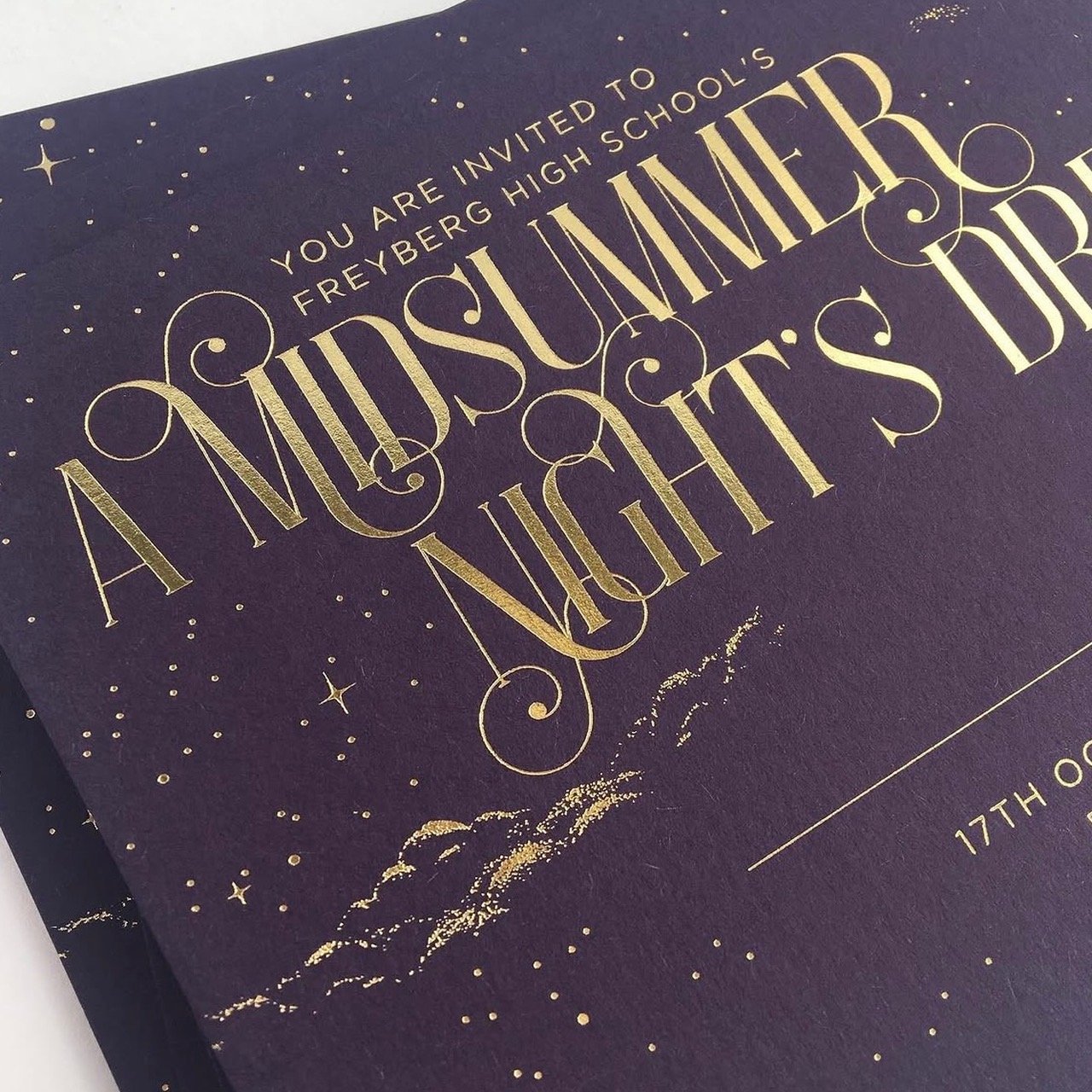 A dark purple invitation card with gold foil lettering that reads 'You are invited to Freyberg High School's A Midsummer Night's Dream Ball' and features illustrations of stars and clouds.