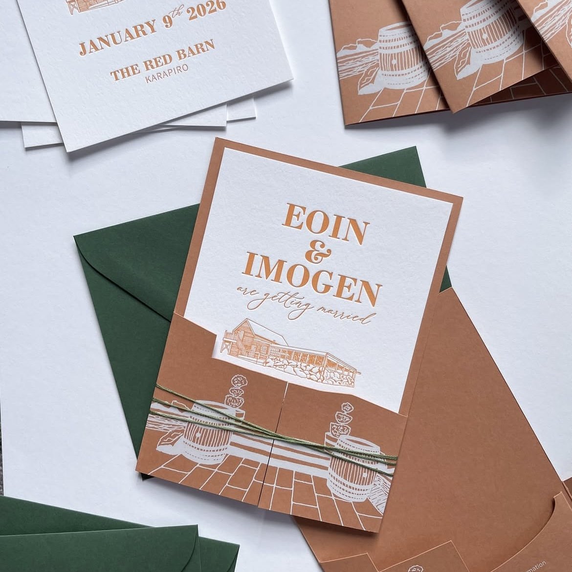 Wedding invitation card with the names EOIN & IMOGEN, indicating they are getting married, with an illustration of a building at the bottom. The card is placed on a green envelope, surrounded by additional cards and paper elements in white, copper, and green colors.