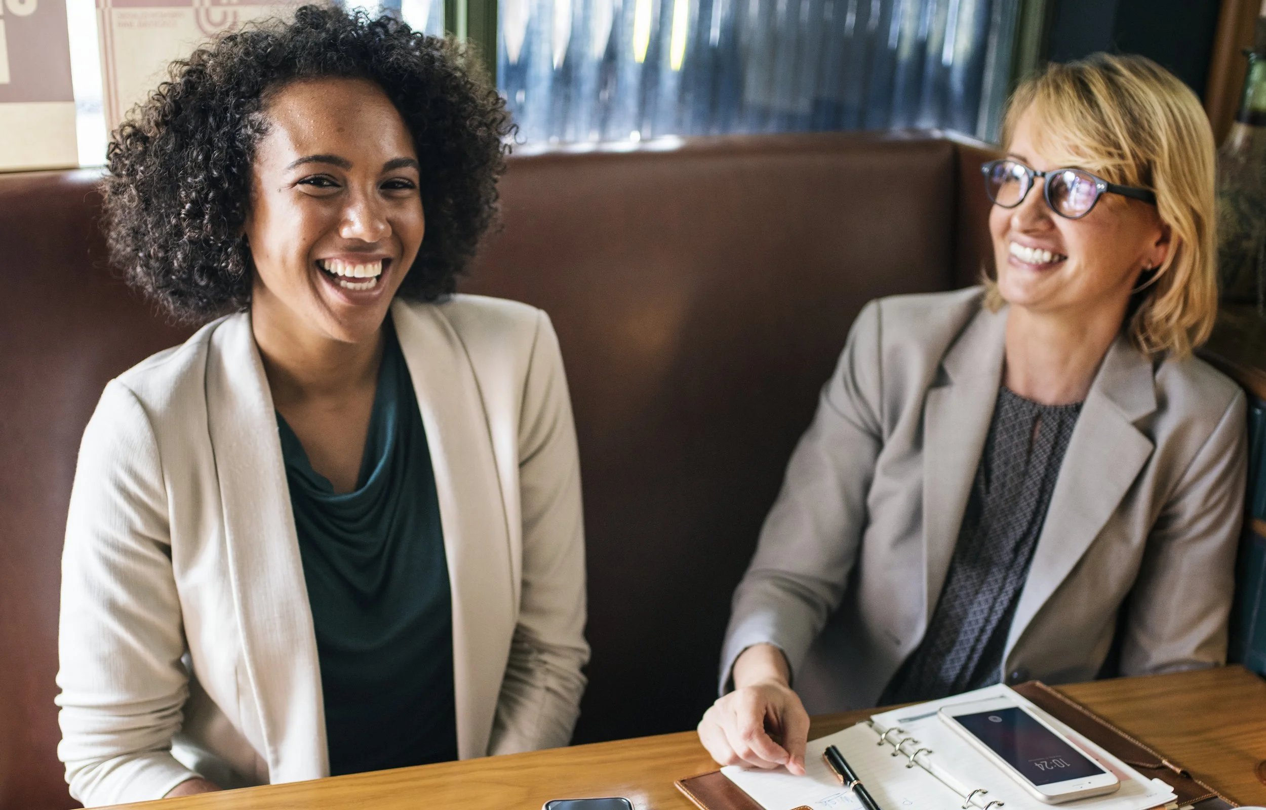 Two women sitting at a table, laughing and smiling during a conversation, in a cozy cafe or restaurant setting.