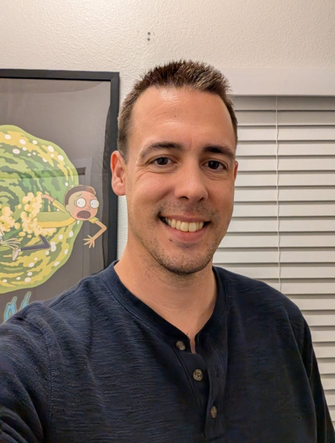 A smiling man taking a selfie indoors next to a framed cartoon poster of a girl running away from a giant yellow-green creature.