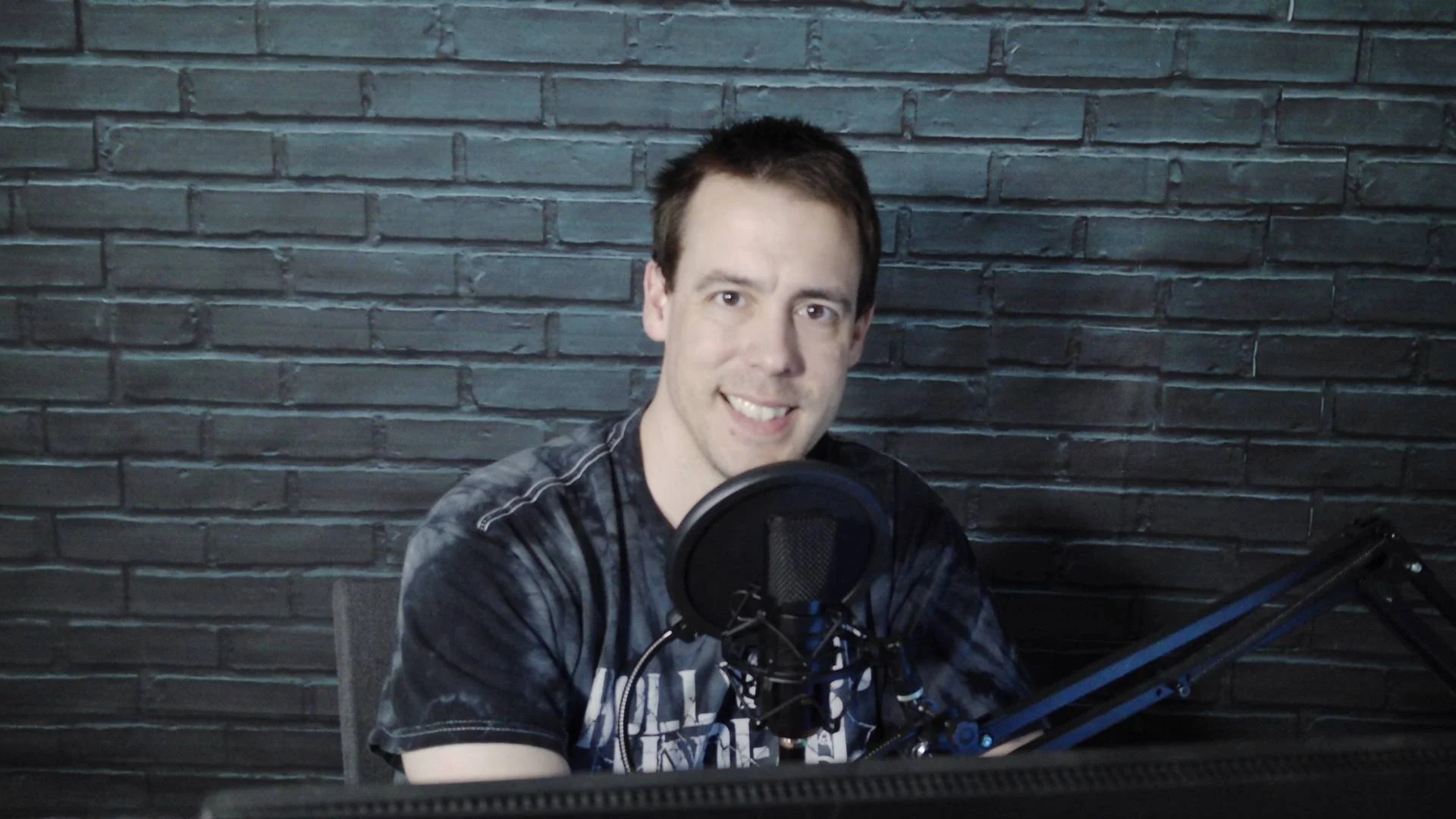 A smiling man with short dark hair in a black T-shirt sits in front of a microphone with a pop filter, against a dark gray brick wall background.
