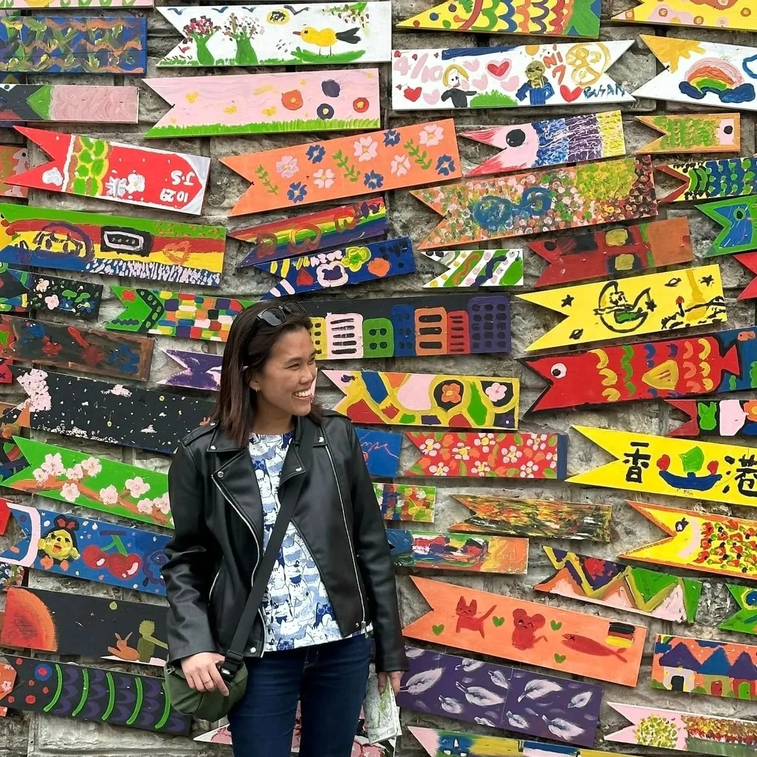 A woman smiling and standing in front of a colorful wall of painted wooden fish shapes, each with different designs and patterns.