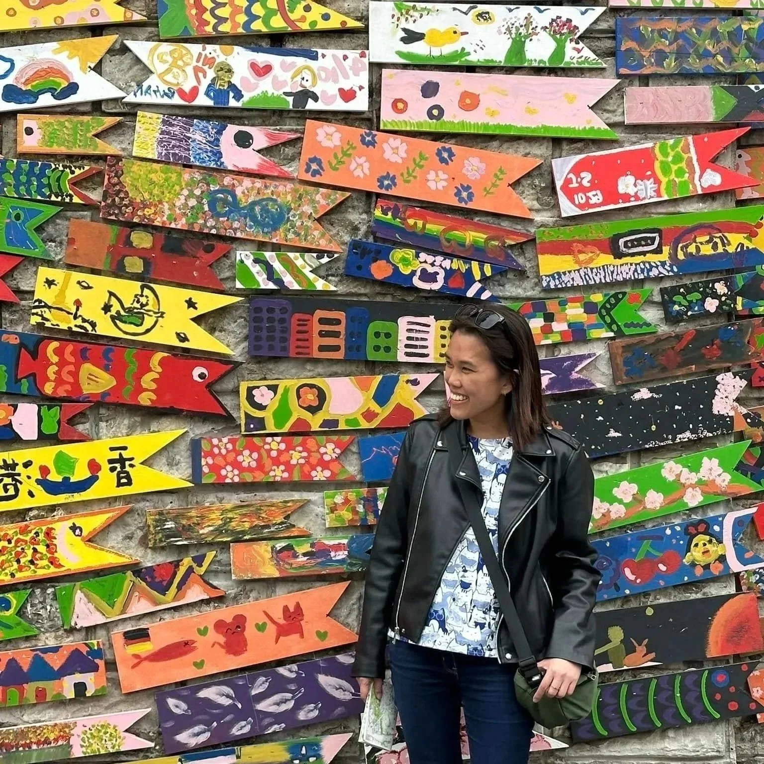 A woman wearing a black leather jacket and jeans standing in front of a colorful wall of painted wooden fish-shaped plaques, smiling and holding paper and a purse.