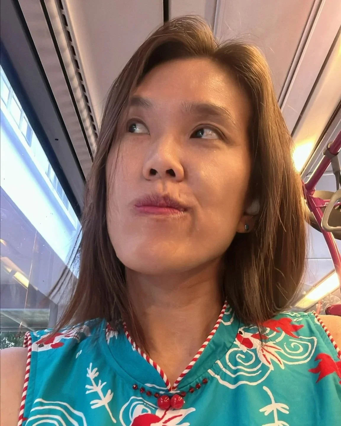 A woman with shoulder-length brown hair wearing a sleeveless blue top with red and white patterns, sitting inside a bus or train.