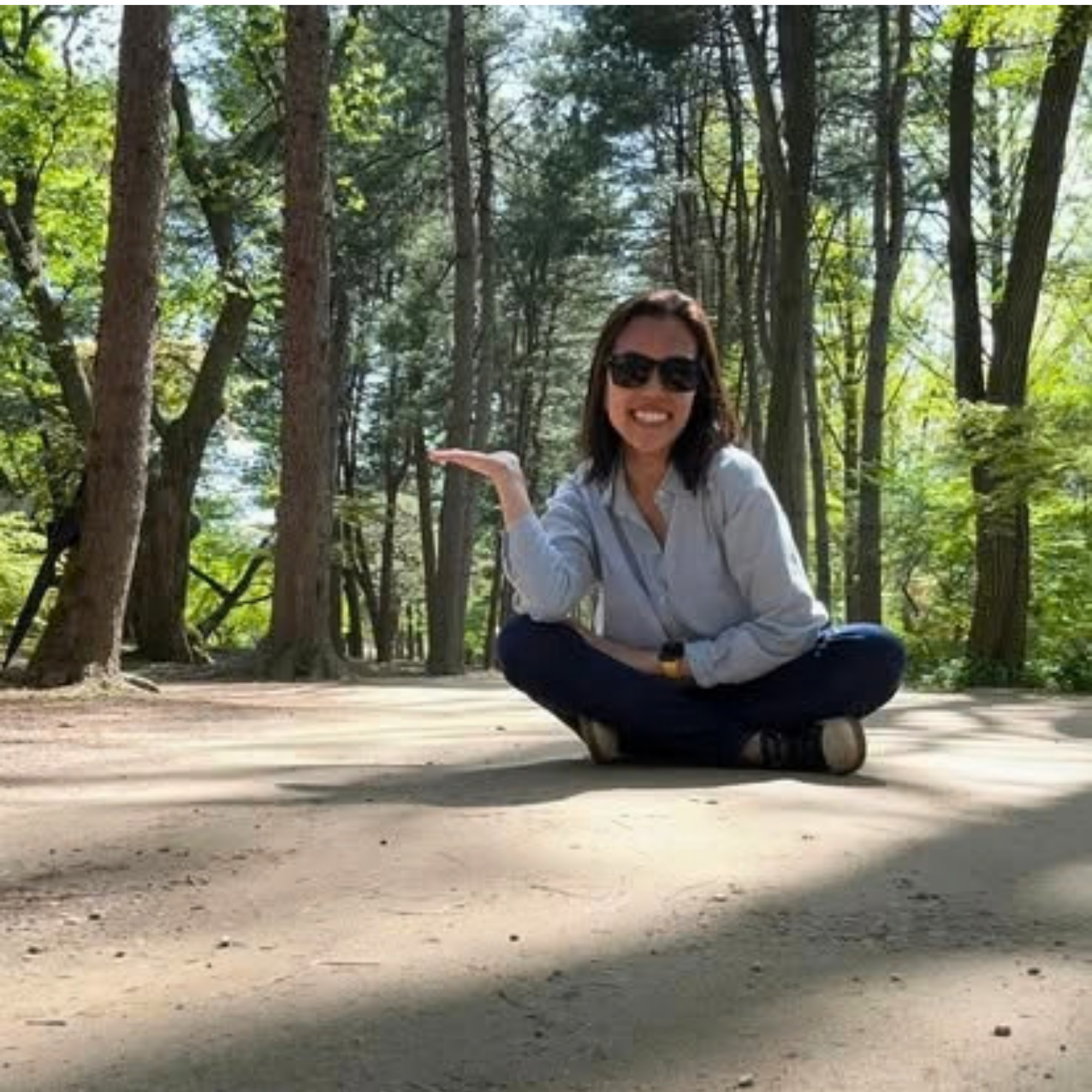 A woman with dark hair and sunglasses sitting cross-legged on the ground in a forest, smiling at the camera while gesturing toward a small skateboard.