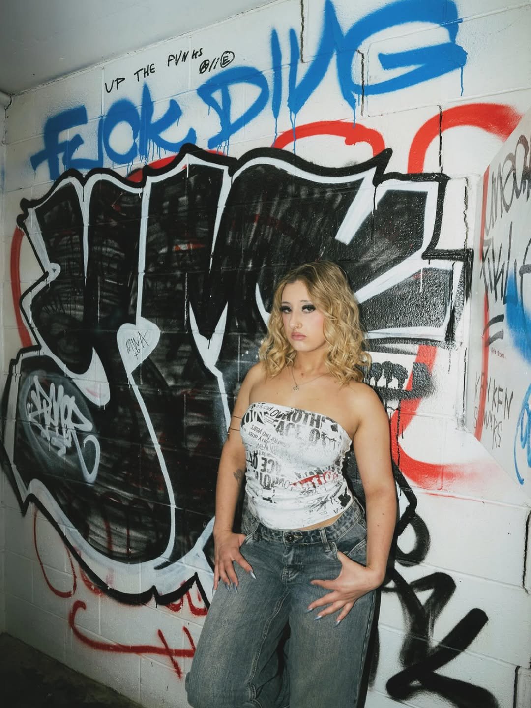 Young woman with blonde curly hair standing in front of graffiti art on a white brick wall, wearing a strapless black and white printed top and loose gray jeans.