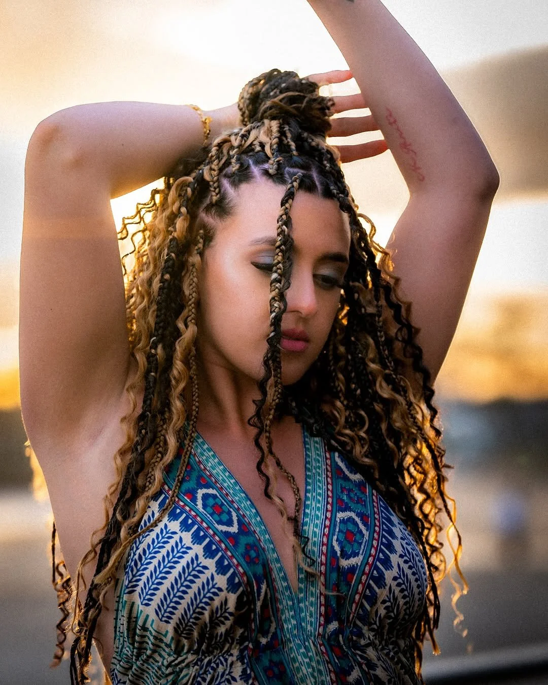 A woman with braided and curly hair, posing with her eyes closed and arms raised above her head, wearing a patterned dress during sunset.
