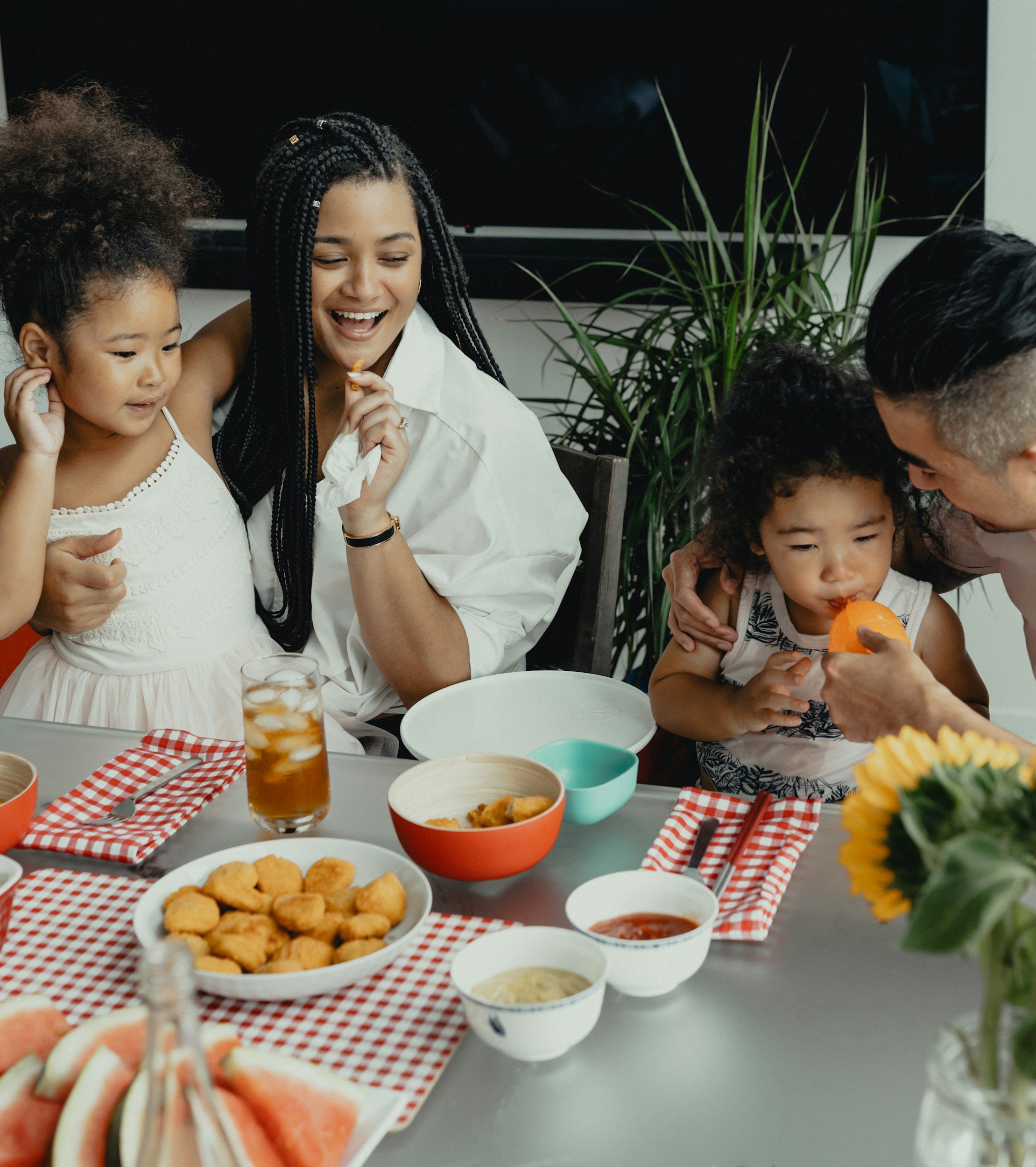 Family gathered around a table with food, including fried chicken, watermelon slices, and dipping sauces, enjoying a meal together.