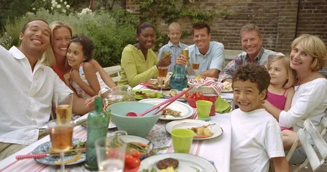 A diverse group of people, including adults and children, gathered around a table outdoors sharing a meal and drinks, smiling and enjoying each other's company.