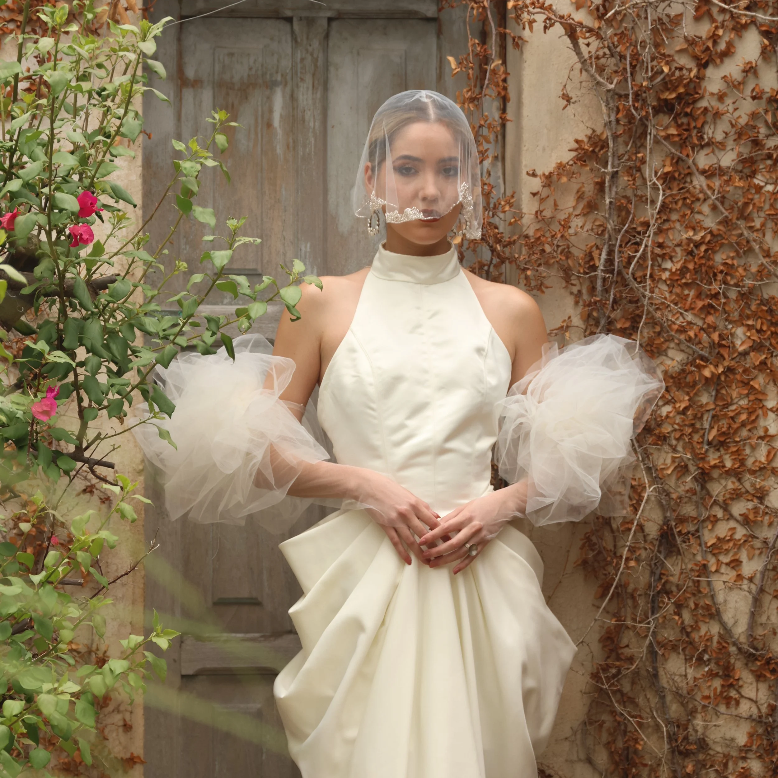 Mujer con vestido de novia color marfil, lleno de volumen, con mangas de tul, lleva velo y pendientes grandes, en un fondo de muro con plantas secas y verdes.