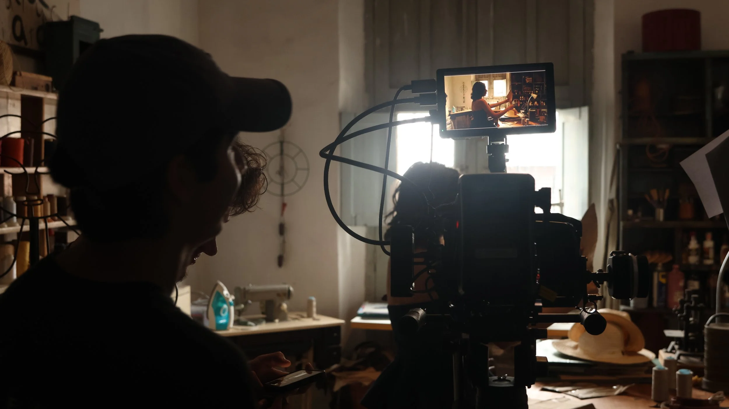 Una escena de grabación en un taller, con una persona operando una cámara y otra revisando en un teléfono móvil, en un ambiente con luz natural y estanterías con herramientas y objetos.