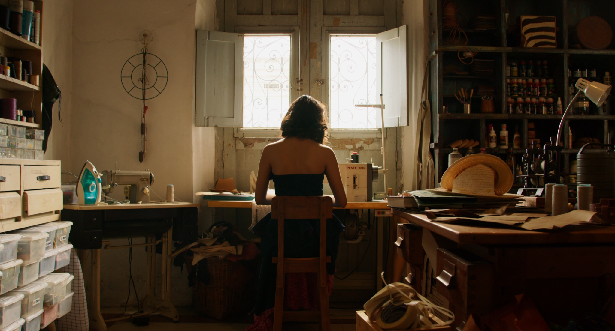 Mujer sentada en una silla trabajando en una máquina de coser en un taller con estanterías llenas de hilos, telas y herramientas de costura, ventanas abiertas que dejan entrar luz natural.