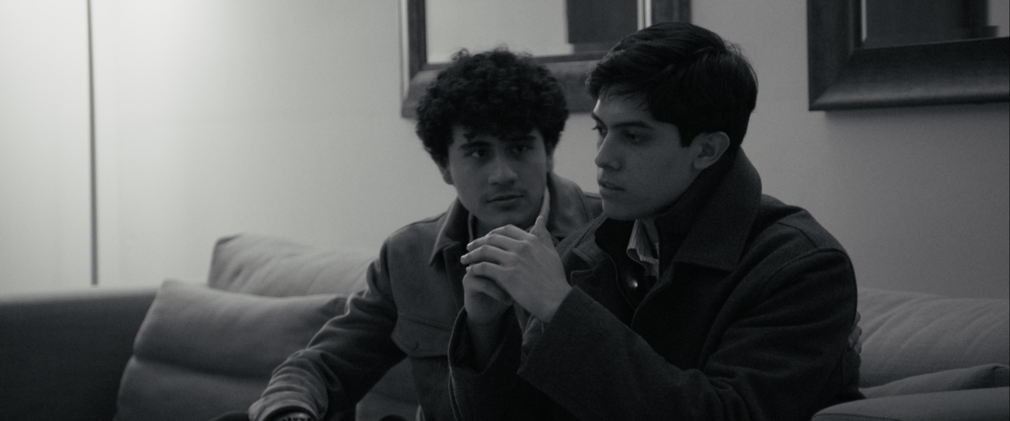 Dos jóvenes sentados en un sofá, uno con cabello rizado y otro con cabello liso, en un ambiente interior, en una escena en tonos grises.