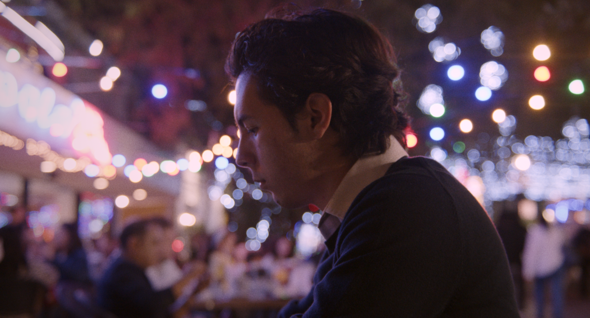 Un joven con cabello oscuro en un lugar con luces de Navidad, viendo hacia abajo, con muchas personas en el fondo en un ambiente festivo nocturno.