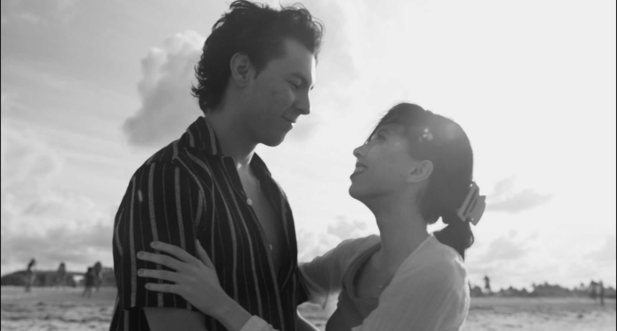 Una pareja en la playa mirando románticamente, ambos sonriendo, con el sol detrás de ellos y el cielo nublado.