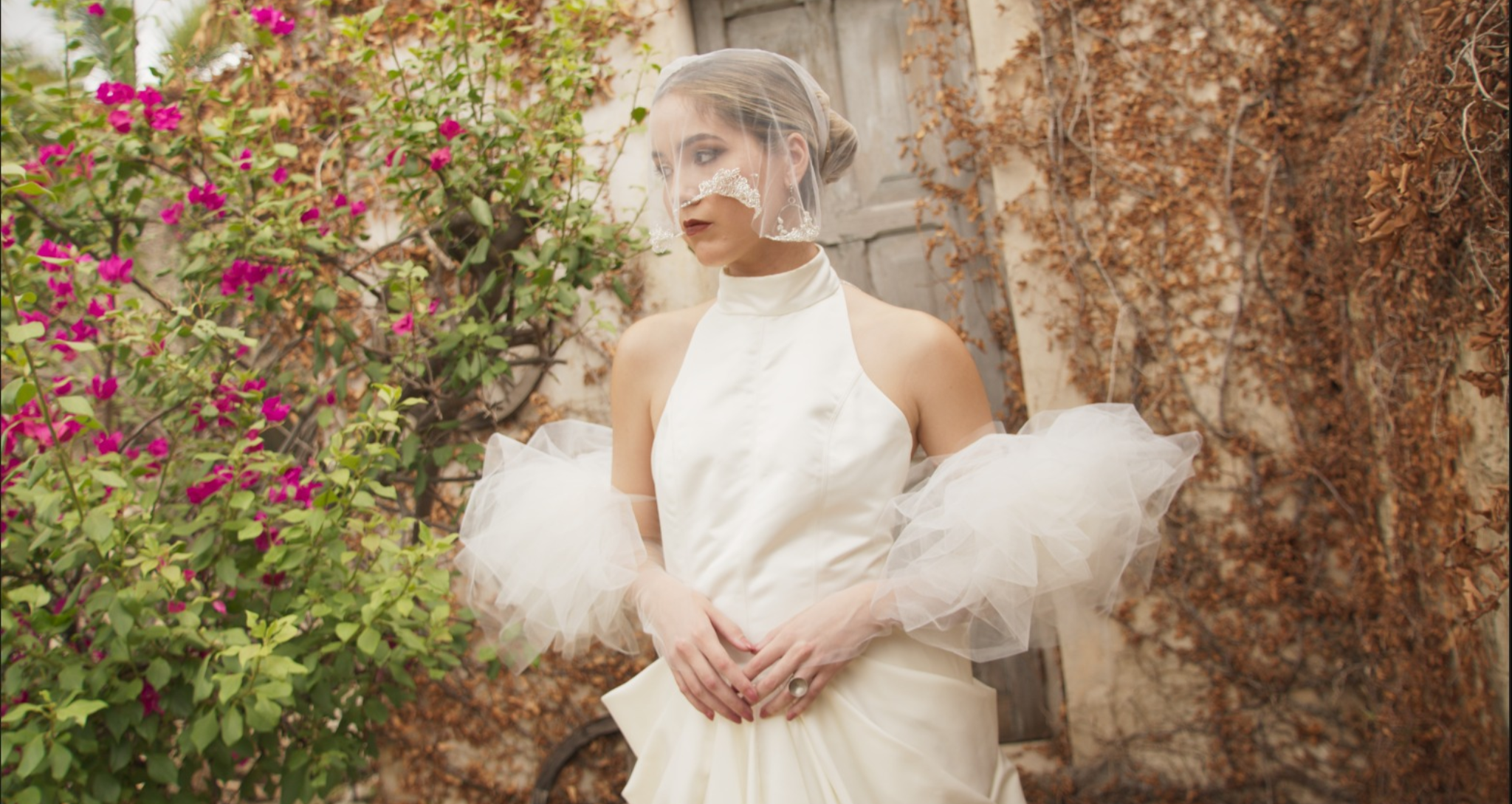 Mujer con vestido de novia y velo de encaje, de perfil, en un jardín con paredes cubiertas de hojas secas y bugs naturales, rodeada de plantas con flores rosadas.