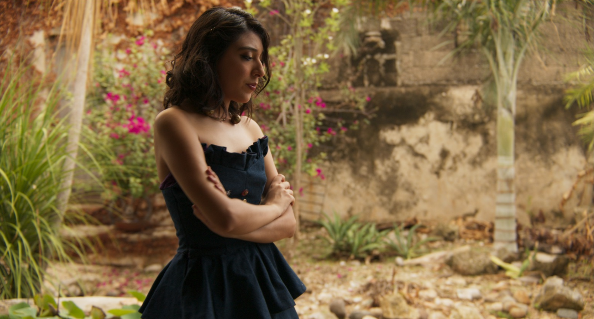 Mujer con vestido oscuro, de cabello rizado, en un jardín con plantas y flores, mirando hacia abajo con los brazos cruzados.