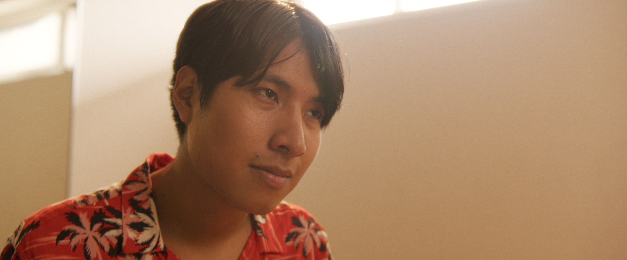 Hombre con cabello oscuro y piel clara, usando una camiseta roja con un patrón de hojas negras y rosas, en un ambiente interior con luz suave y fondo nublado beige.