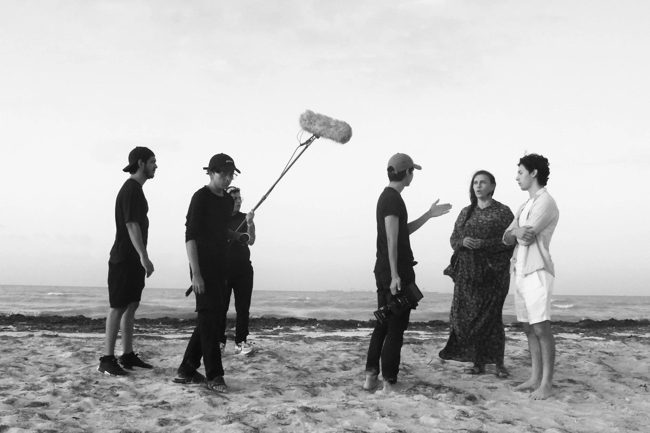 Equipo de producción filmando una escena en la playa con personas hablando y un micrófono de boom en negro y blanco.