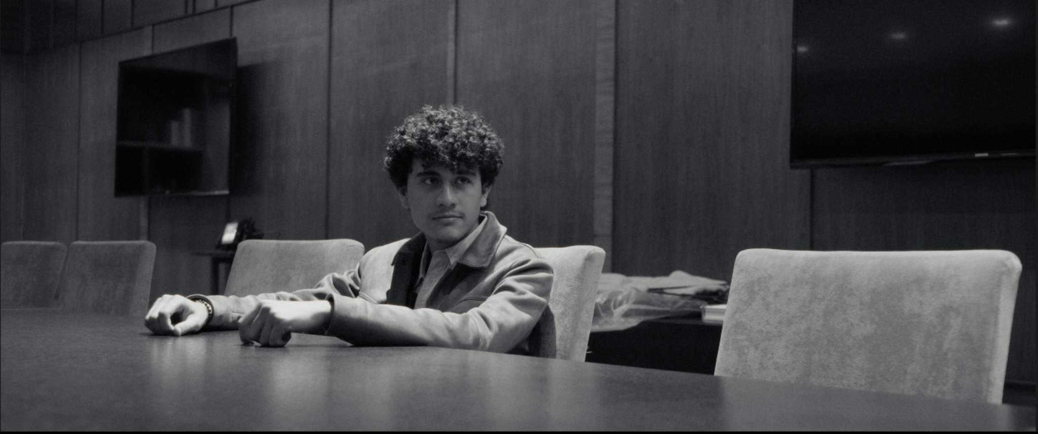 Joven con cabello rizado sentado en una sala de reuniones con paredes de madera, sillas y mesa larga, en un ambiente profesional en blanco y negro.