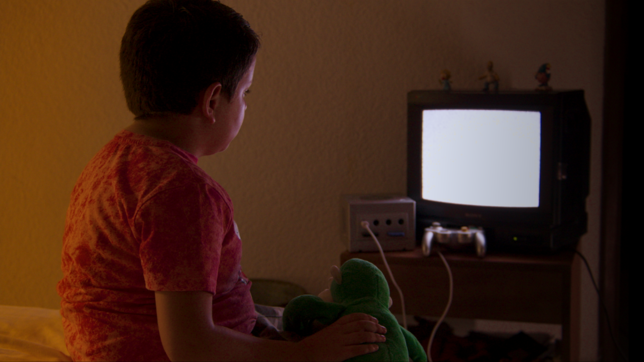 Un niño sentado en la cama sosteniendo un muñeco de peluche verde, mirando una televisión vieja encendida en una habitación oscura.