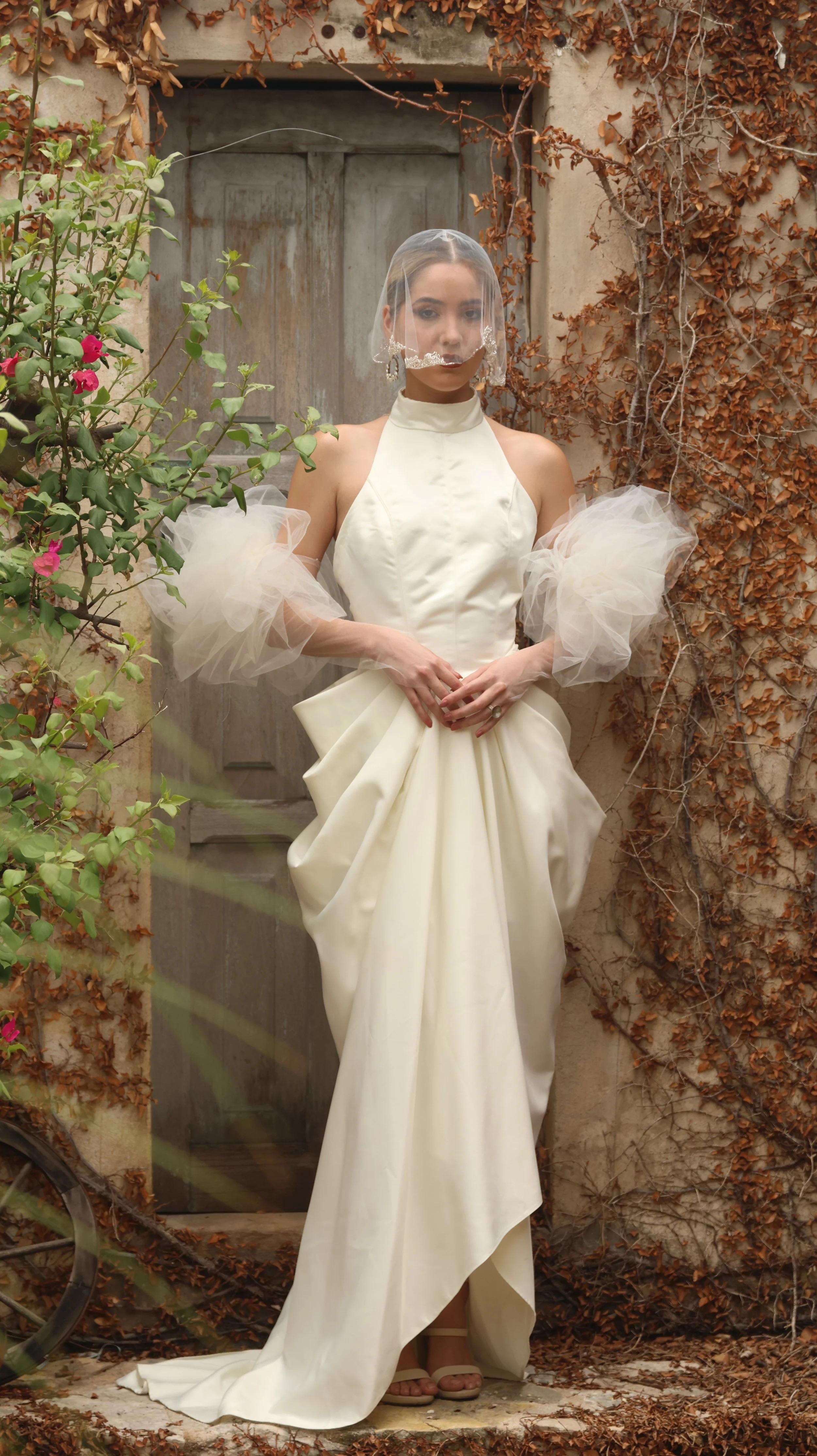 Mujer con vestido de novia y velo, posando frente a una puerta de madera antigua rodeada de plantas.