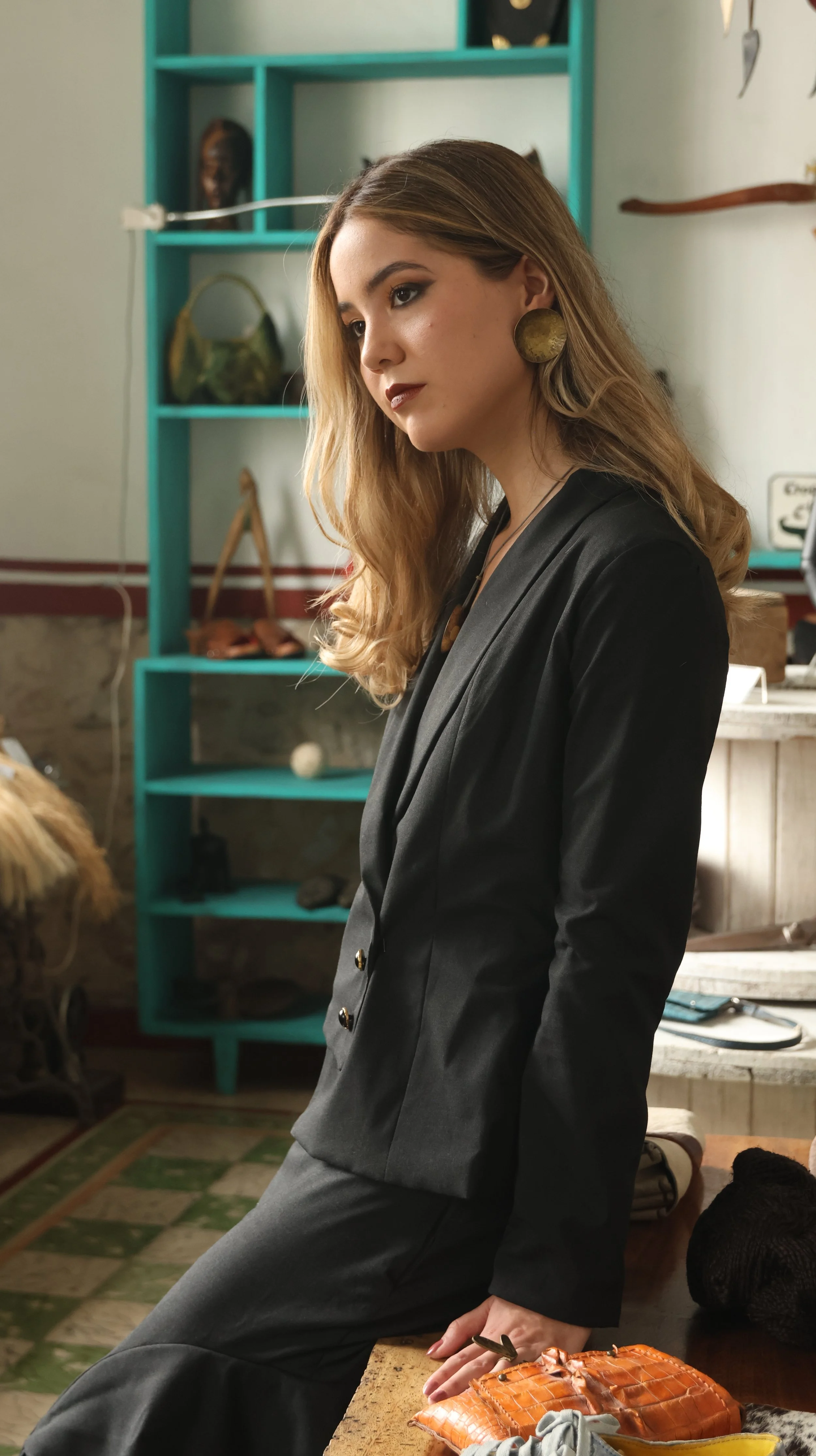 Mujer con cabello rubio y ondulado, vestida con saco negro y pantalones oscuros, apoyada en una mesa de madera, en un entorno que parece ser una tienda o una sala decorada con objetos artesanales y decorativos.