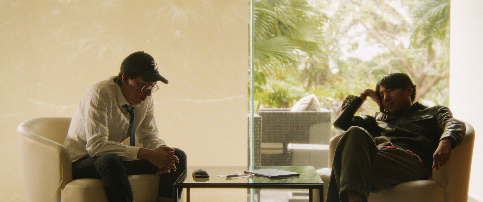 Dos jóvenes sentados en sillones en una sala con vista a un jardín, uno parece preocupado y el otro relajado.