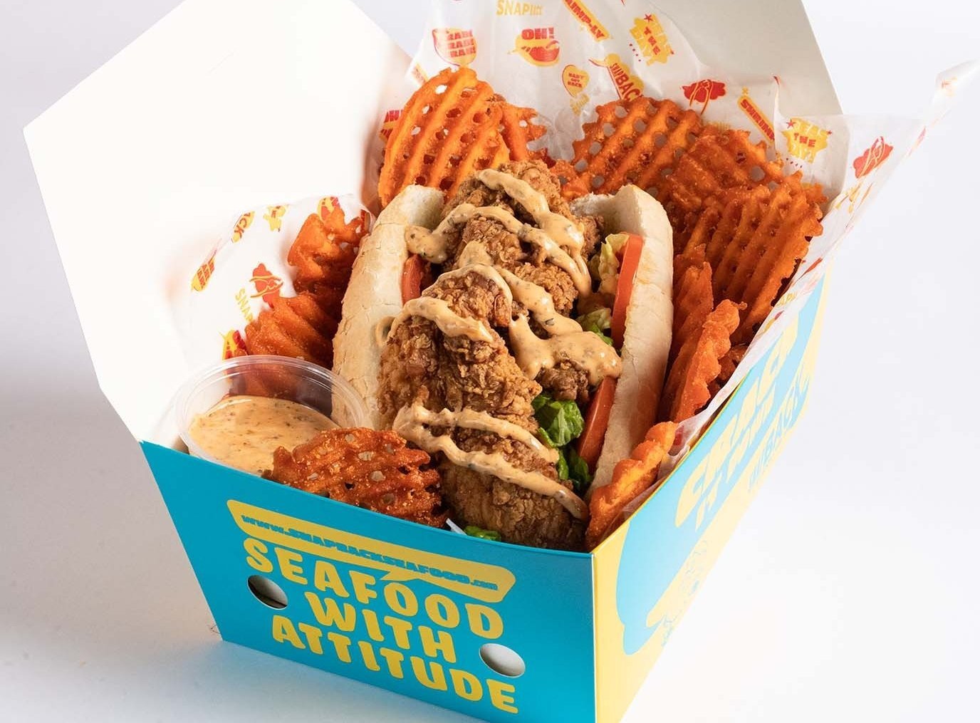 A box of fast food containing fried chicken po'boy sandwich topped with sauce, waffle fries, a potato waffle fry, and a small container of dipping sauce.