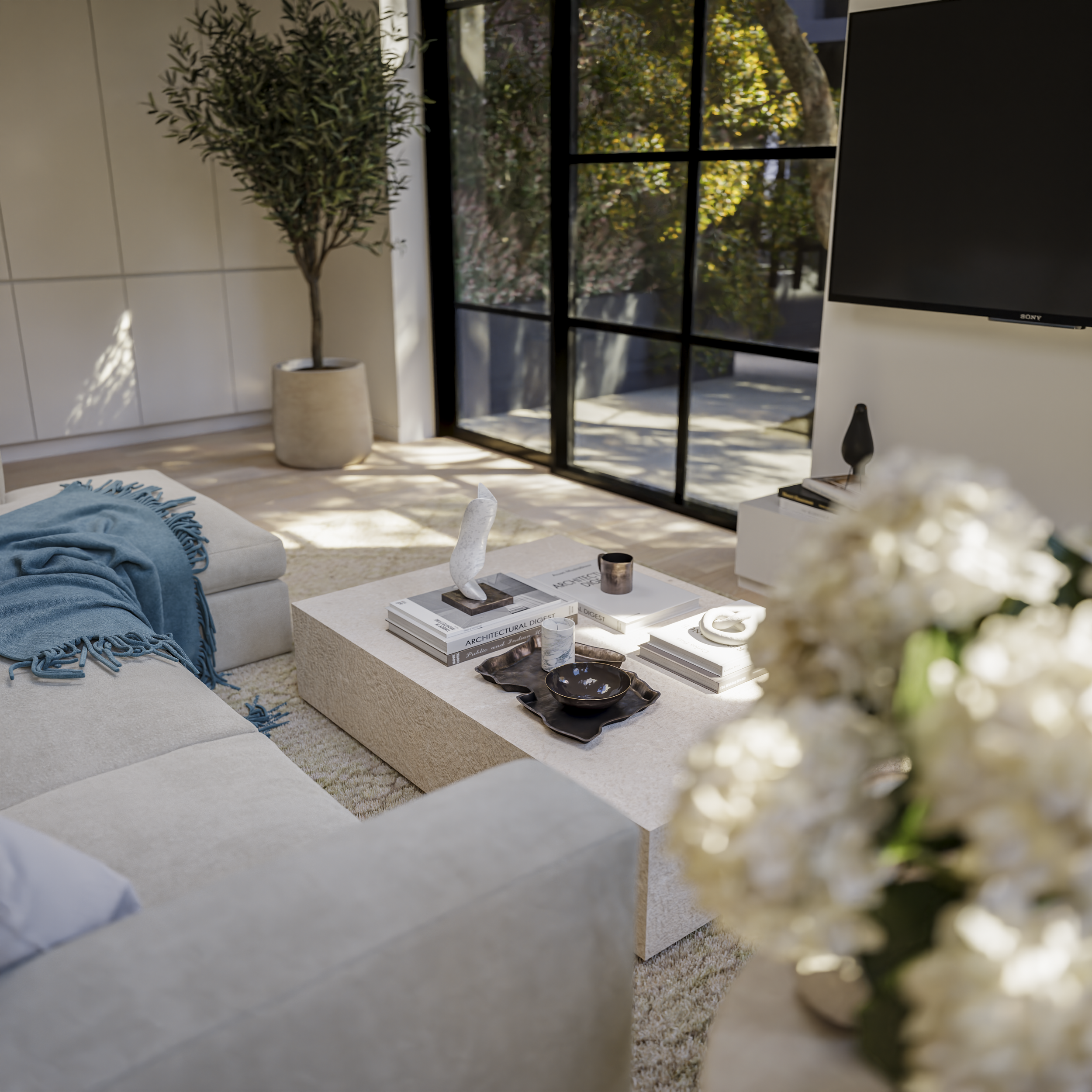 Living room with a beige sofa, a coffee table with books and decorative items, a large potted plant, a wall-mounted TV, and glass sliding doors with a view of trees outside.