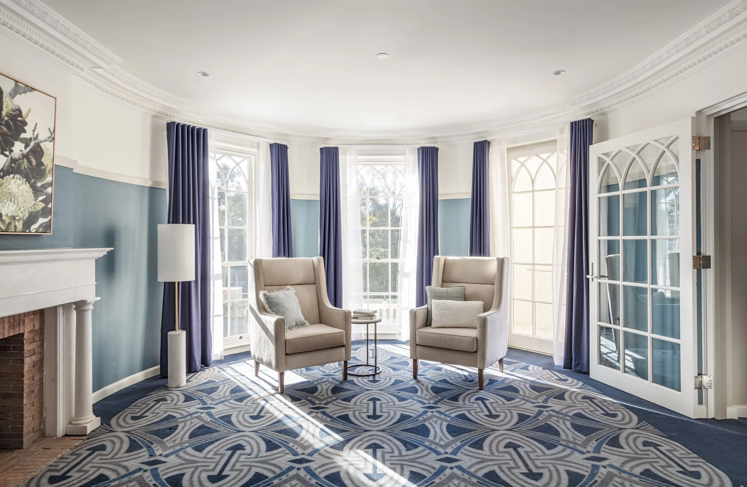 Bright sitting area with two beige armchairs and pillows, in front of large windows with white and navy curtains, white door with glass panels, and a decorative blue patterned carpet, in a well-lit room.