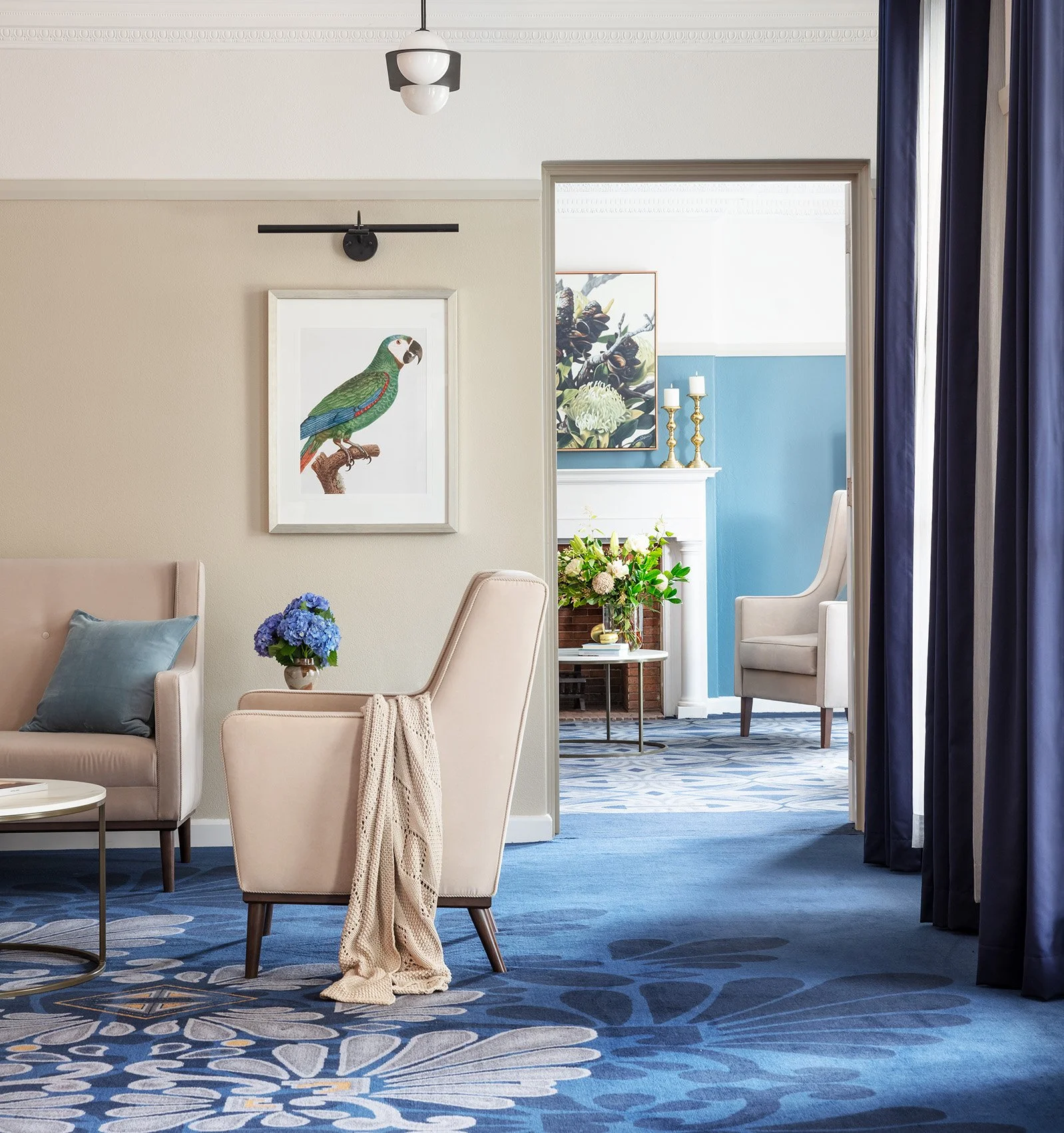 A contemporary living room with beige and blue decor, including a beige sofa with blue pillows, a beige armchair with a blanket, and a blue floral rug. There are fresh flowers on tables and framed bird artwork on the wall. Curtains are blue, and a doorway leads to a bright room with more floral decorations and artwork.