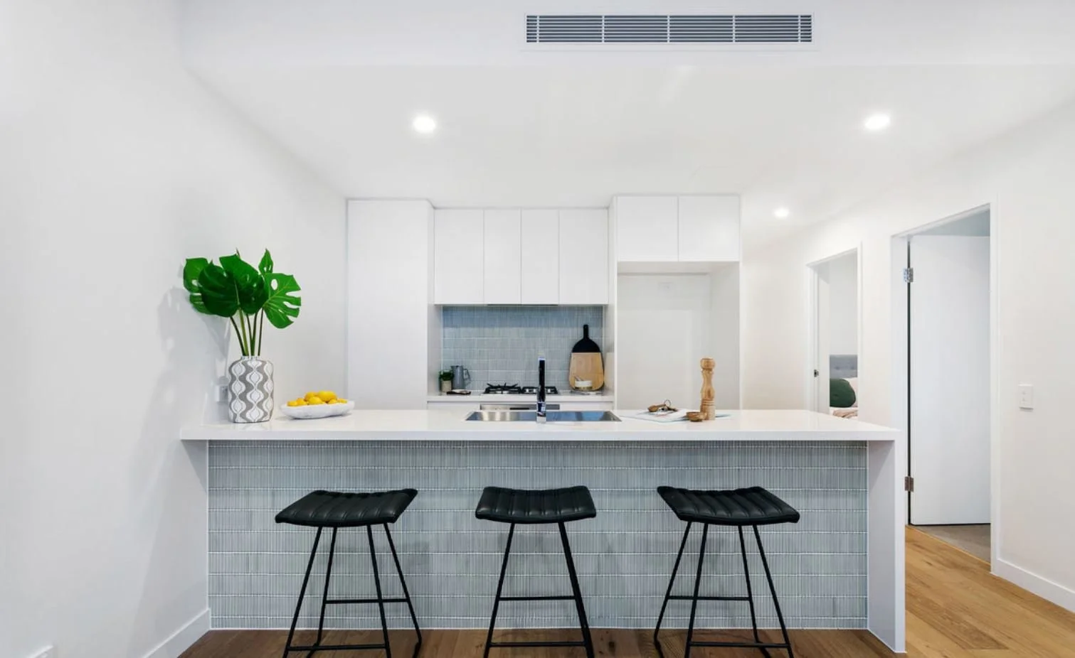 Minimalist kitchen with white cabinets, gray backsplash, and black bar stools at a white breakfast bar with a tiled front. Decor includes large green leaves in a vase and a bowl of lemons.