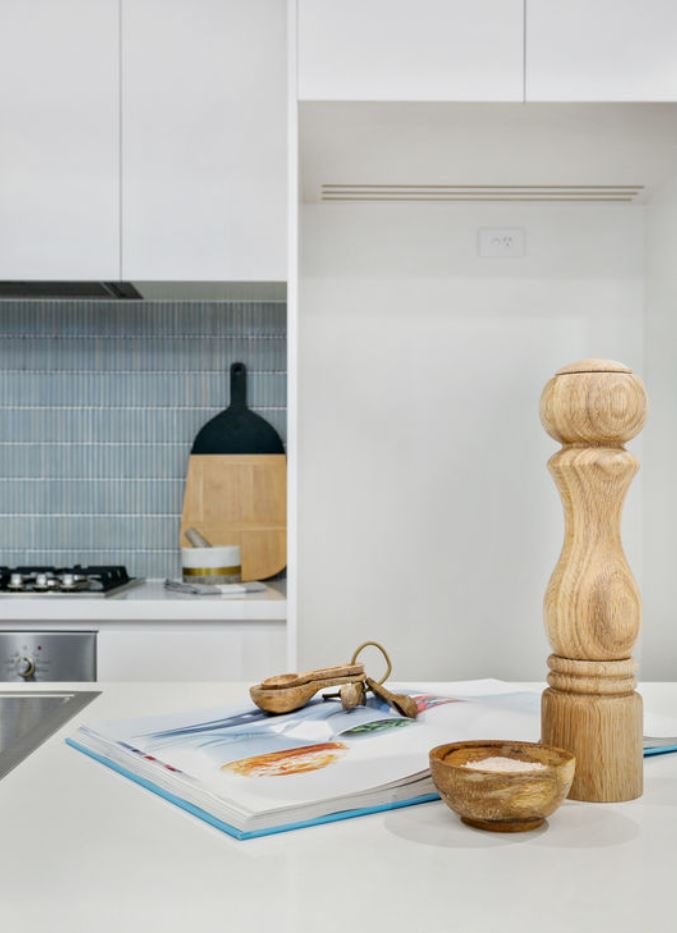 Kitchen counter with garlic, a cookbook, wooden bowls, a wooden salt or pepper mill, and a small spoon, with kitchen cabinets and a backsplash in the background.