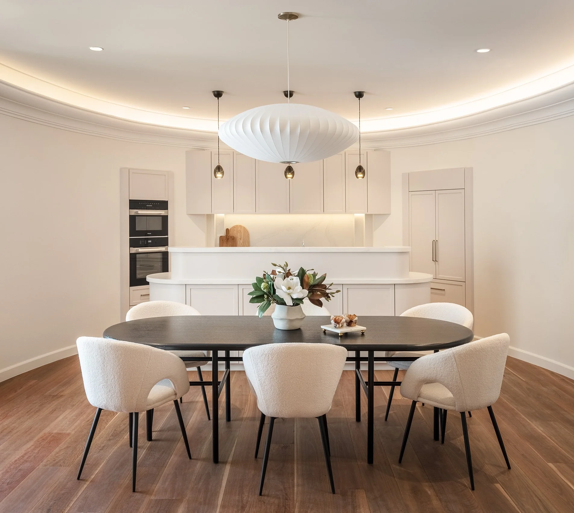 Modern dining area with a dark oval table surrounded by six white upholstered chairs, a vase of white flowers on the table, and a white kitchen in the background with cabinetry and built-in appliances. A large white pendant light hangs above the table, and the room features warm wood flooring.