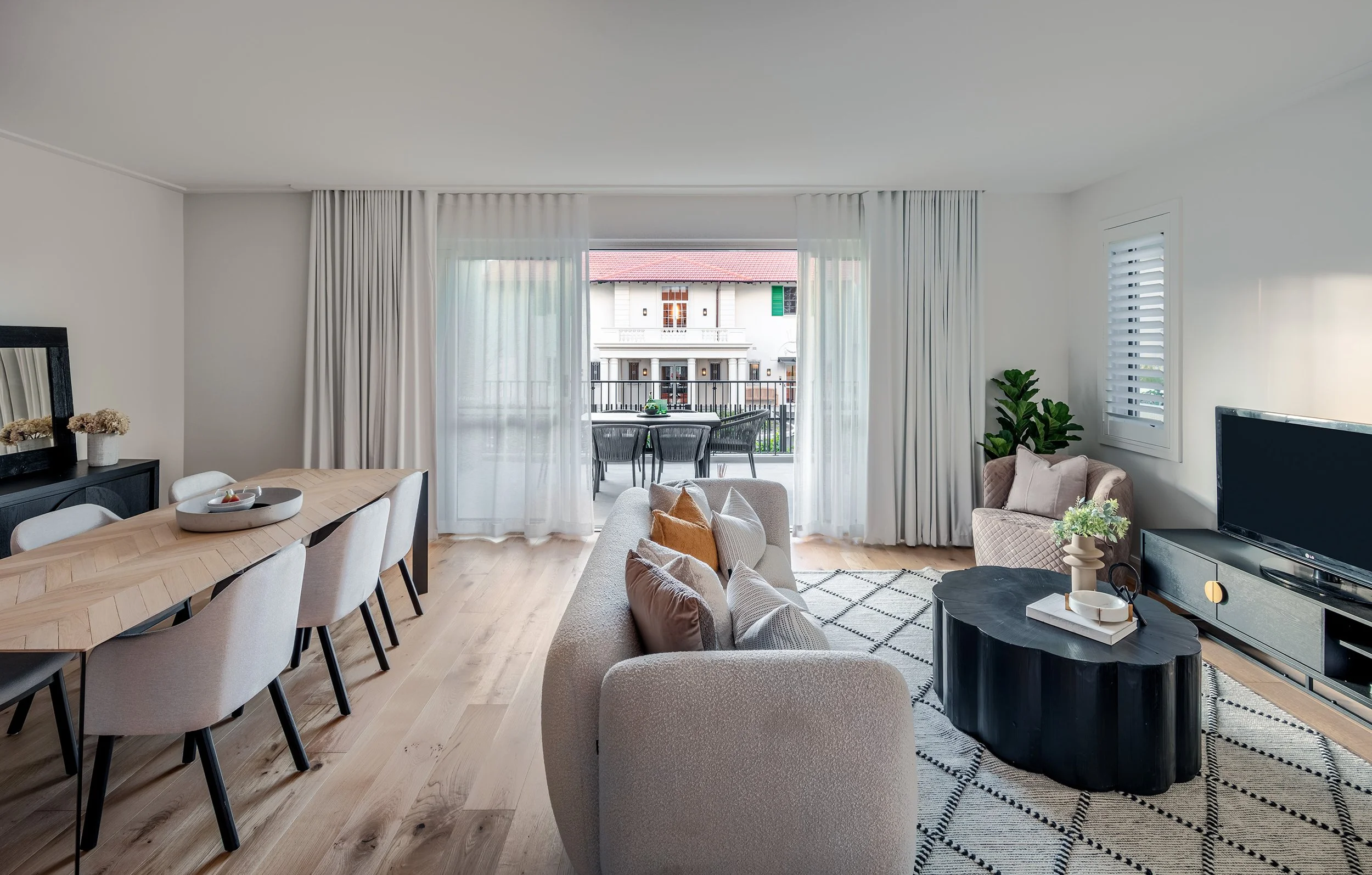 Living room within a luxury retirement living unit with a beige sofa and pillows, a black coffee table with decorative items, a TV on a black console, a dining table with chairs, and sliding glass doors leading to a balcony with outdoor seating.