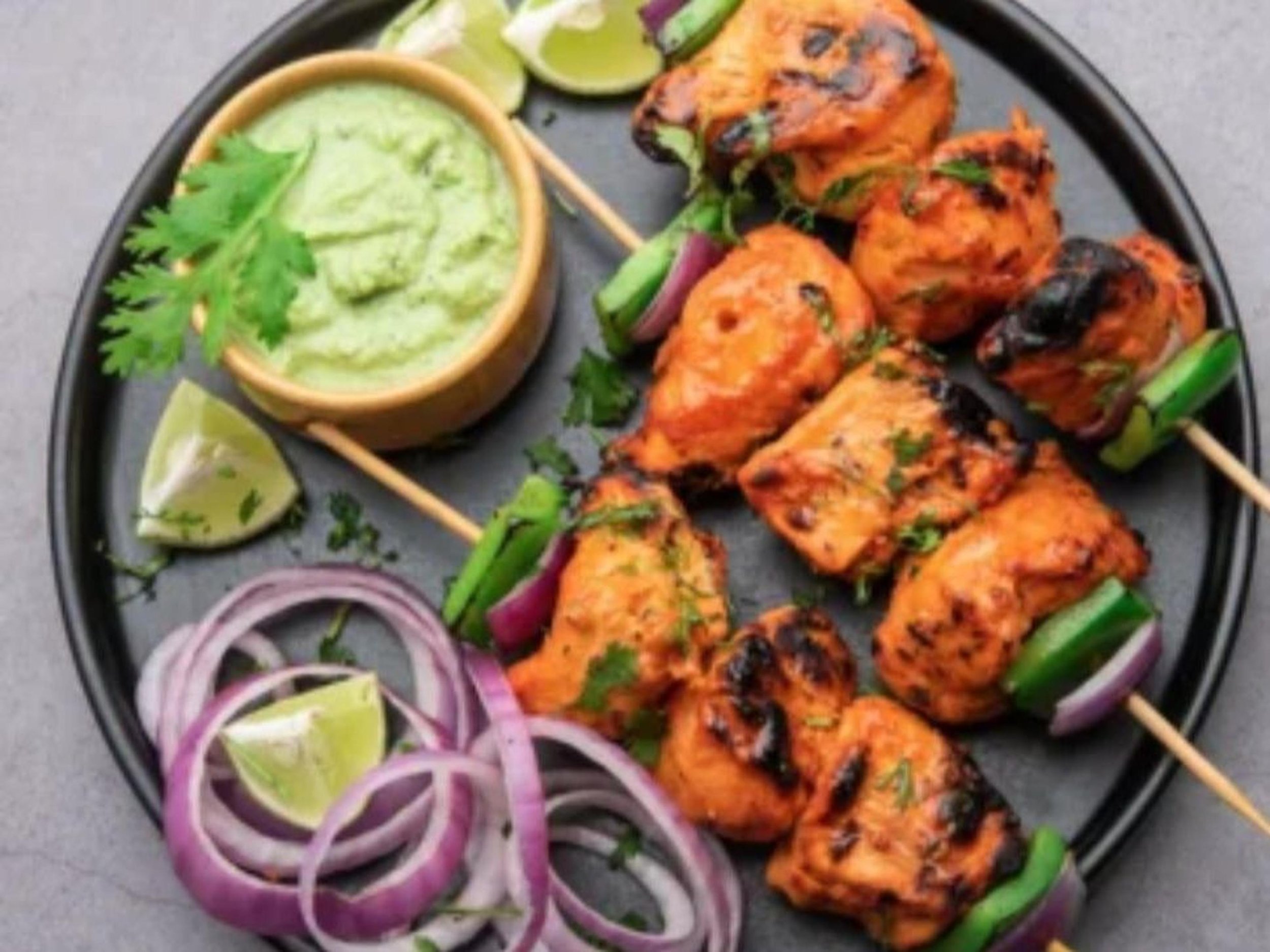 Plate of grilled chicken skewers with green peppers and onions, served with sliced red onions, lime wedges, and a side of green chutney.