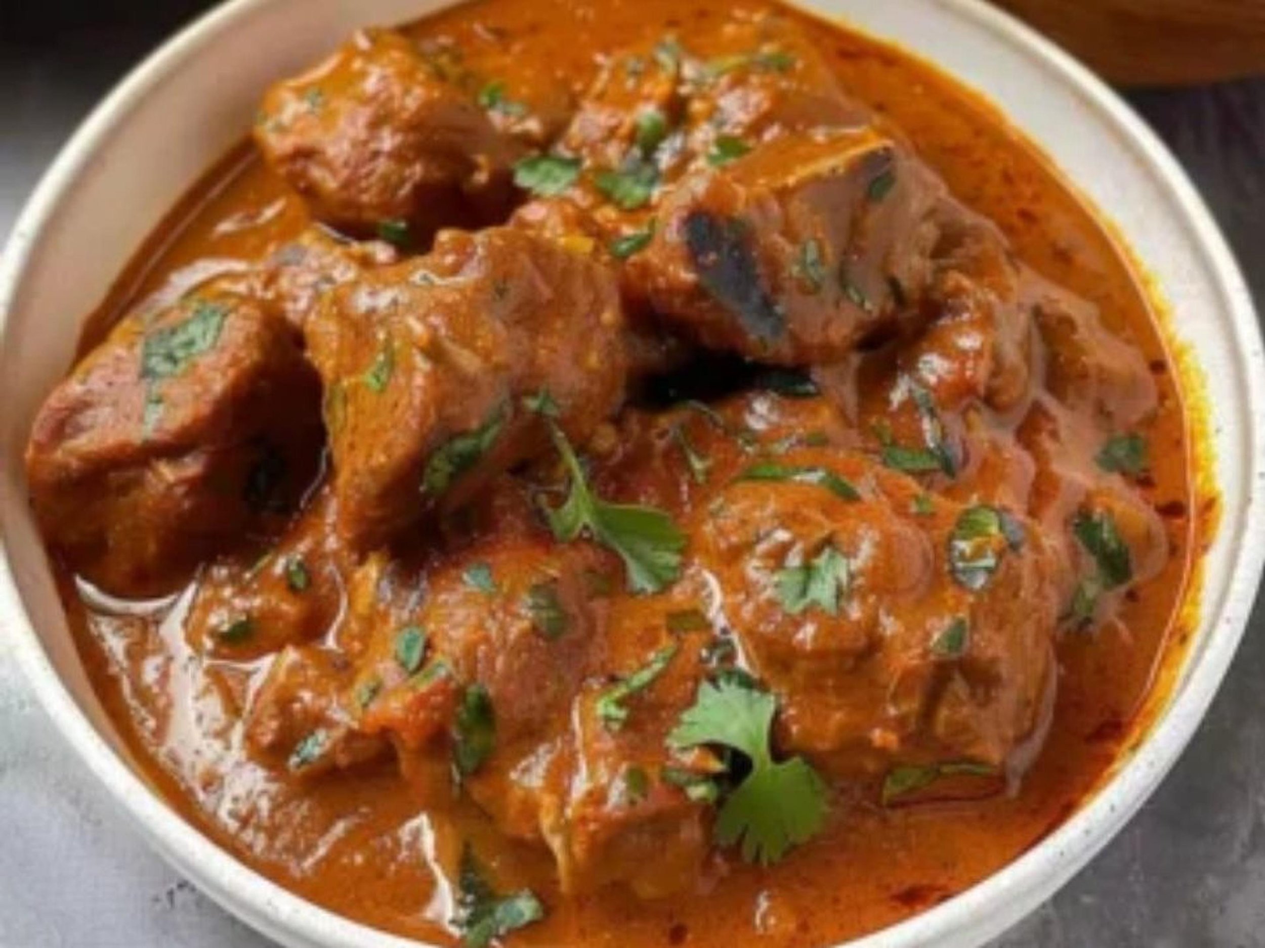 A bowl of Indian chicken curry garnished with cilantro, in a rich, reddish-orange sauce.