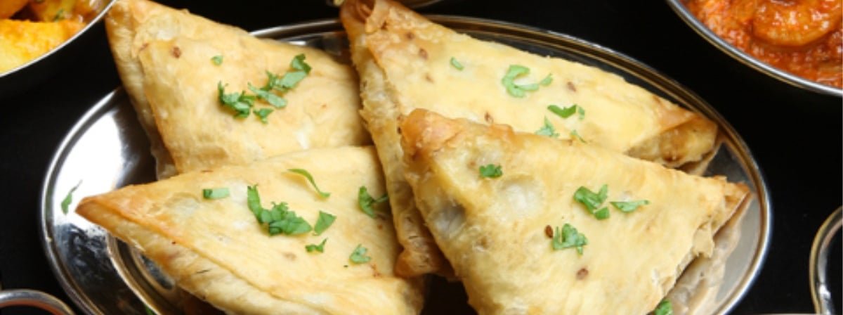 Close-up of four samosas garnished with chopped cilantro, served on a stainless steel plate, with bowls of curry and other dishes partially visible around.
