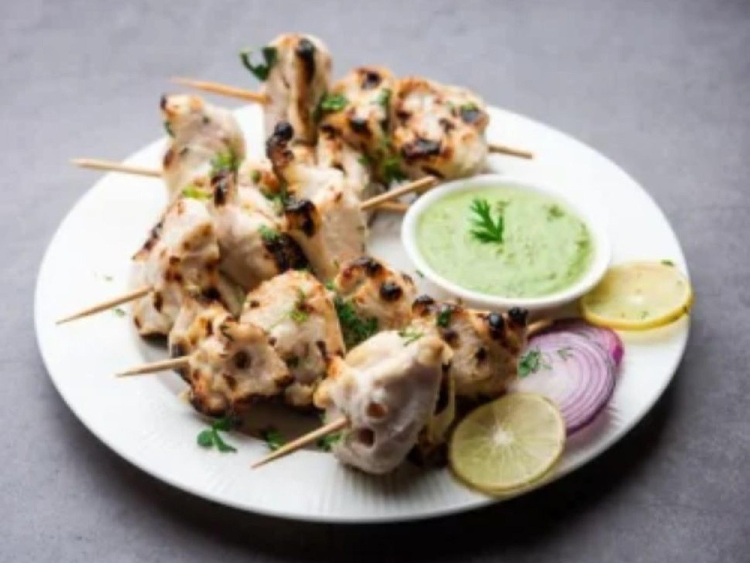 Plate of grilled chicken kebabs garnished with cilantro, served with lemon slices, sliced red onions, and a small bowl of green dipping sauce garnished with cilantro.