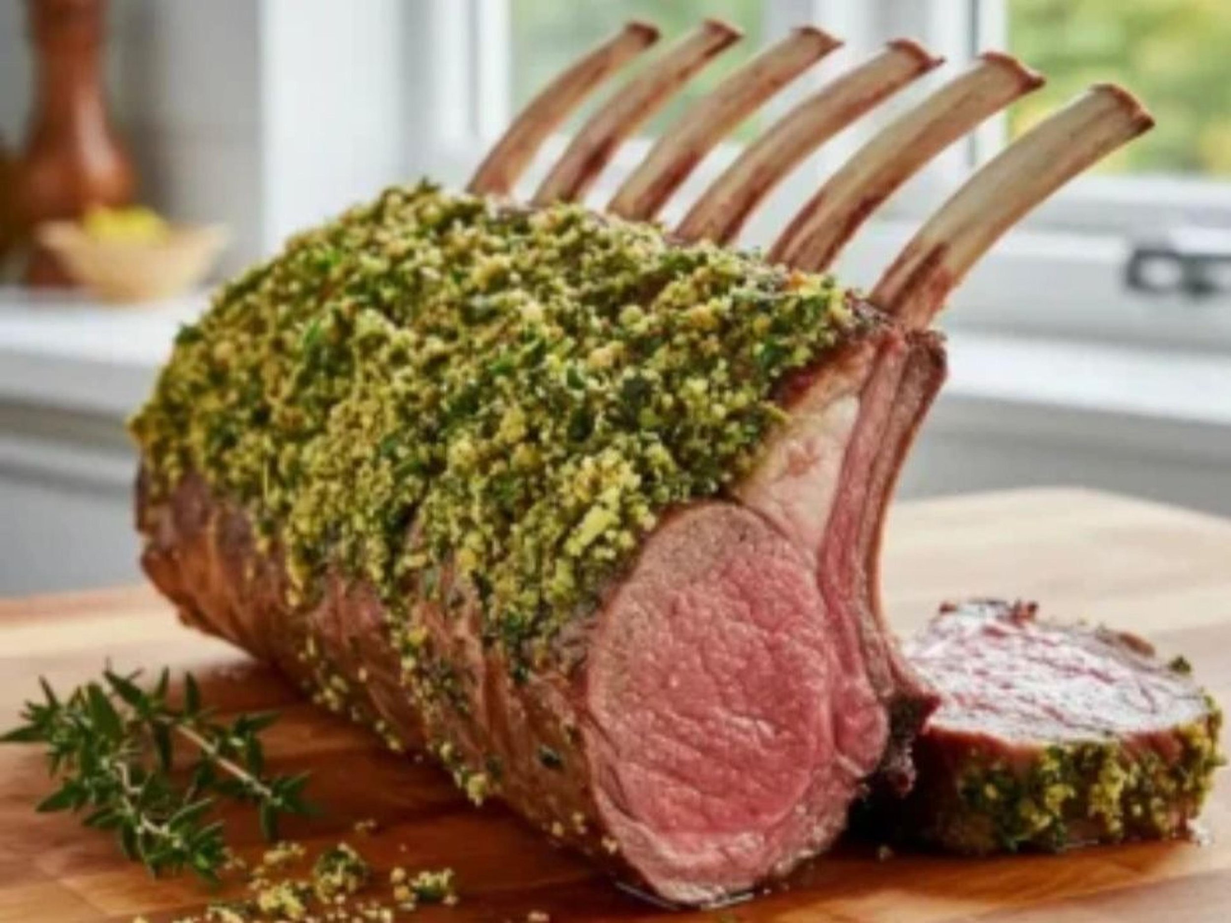 A roasted rack of lamb with herb crust on a wooden cutting board, with one slice cut and placed beside it.