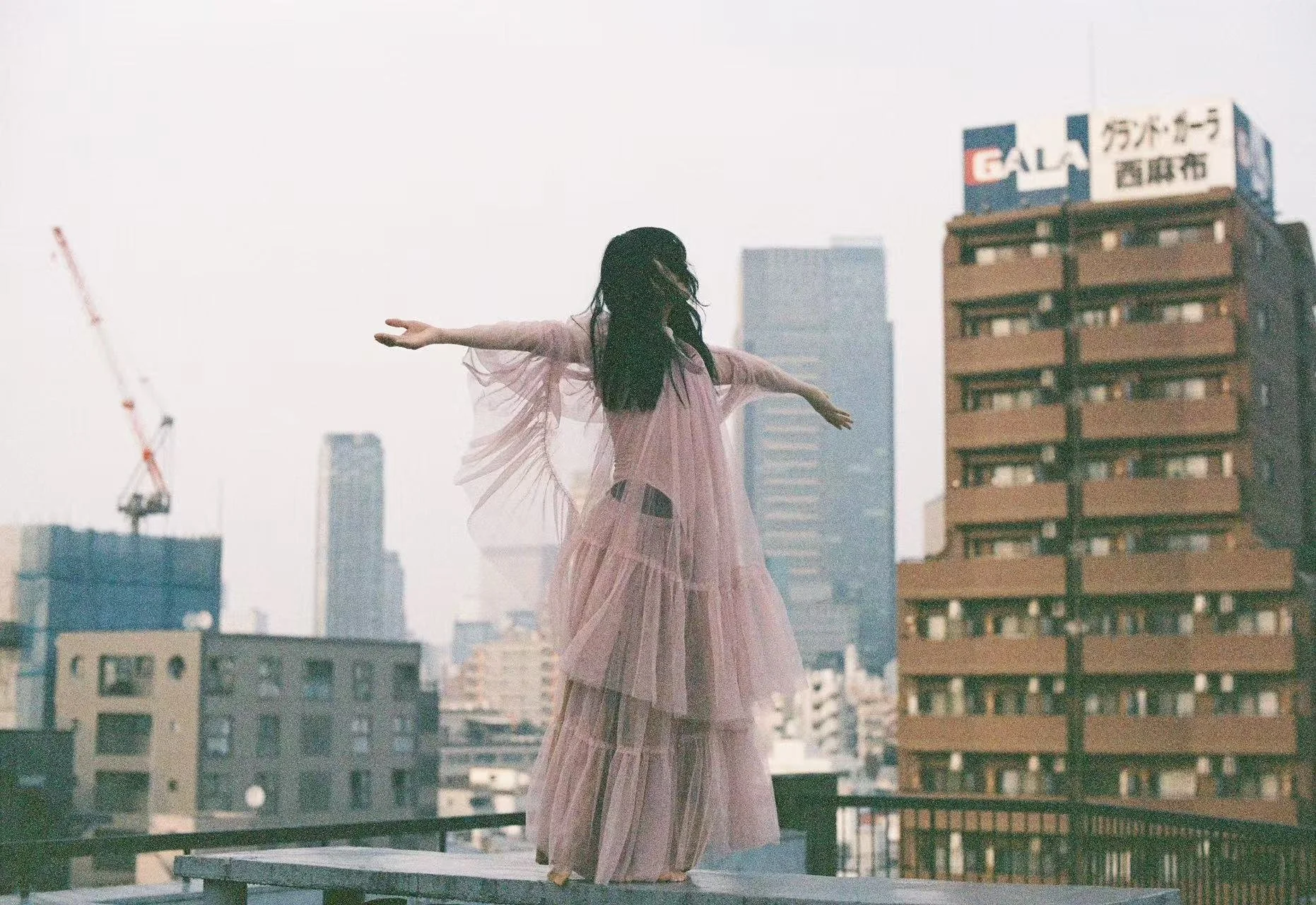 People participating in a rooftop yoga or meditation session in an urban city with tall buildings in the background. A woman in a pink dress is seen standing on a platform, with others sitting or standing nearby.