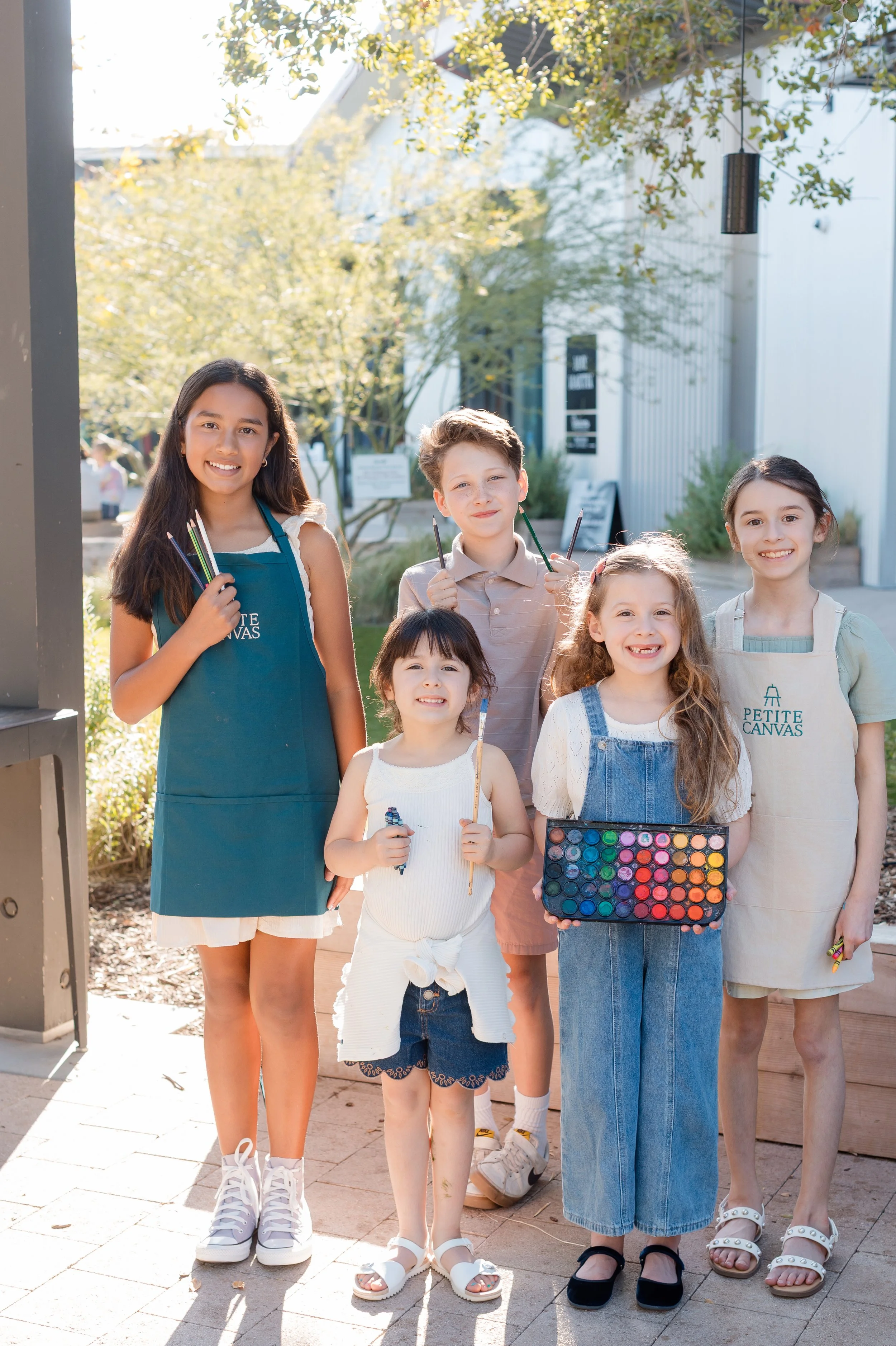 Group of five young artists smiling outdoors while holding paintbrushes, colored pencils, and a watercolor palette.