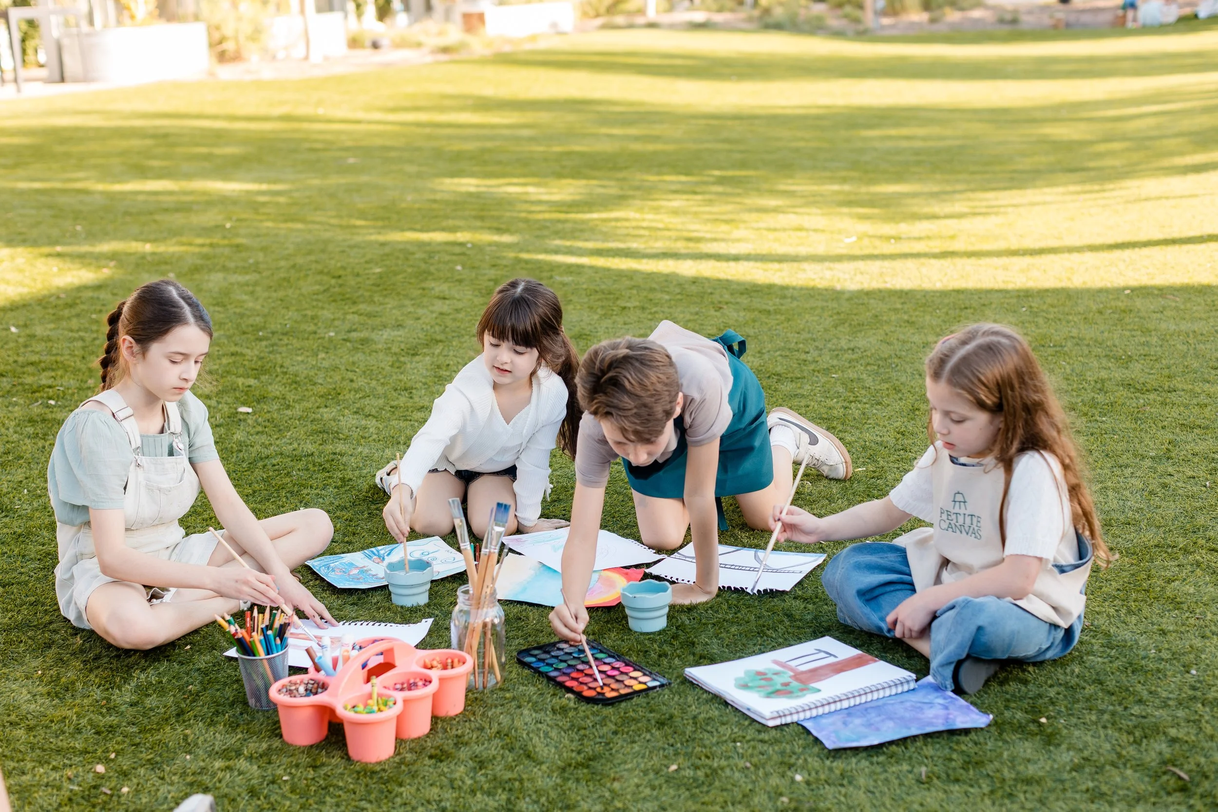 Four young artists painting and drawing together outdoors with watercolor paints, brushes, and art supplies.