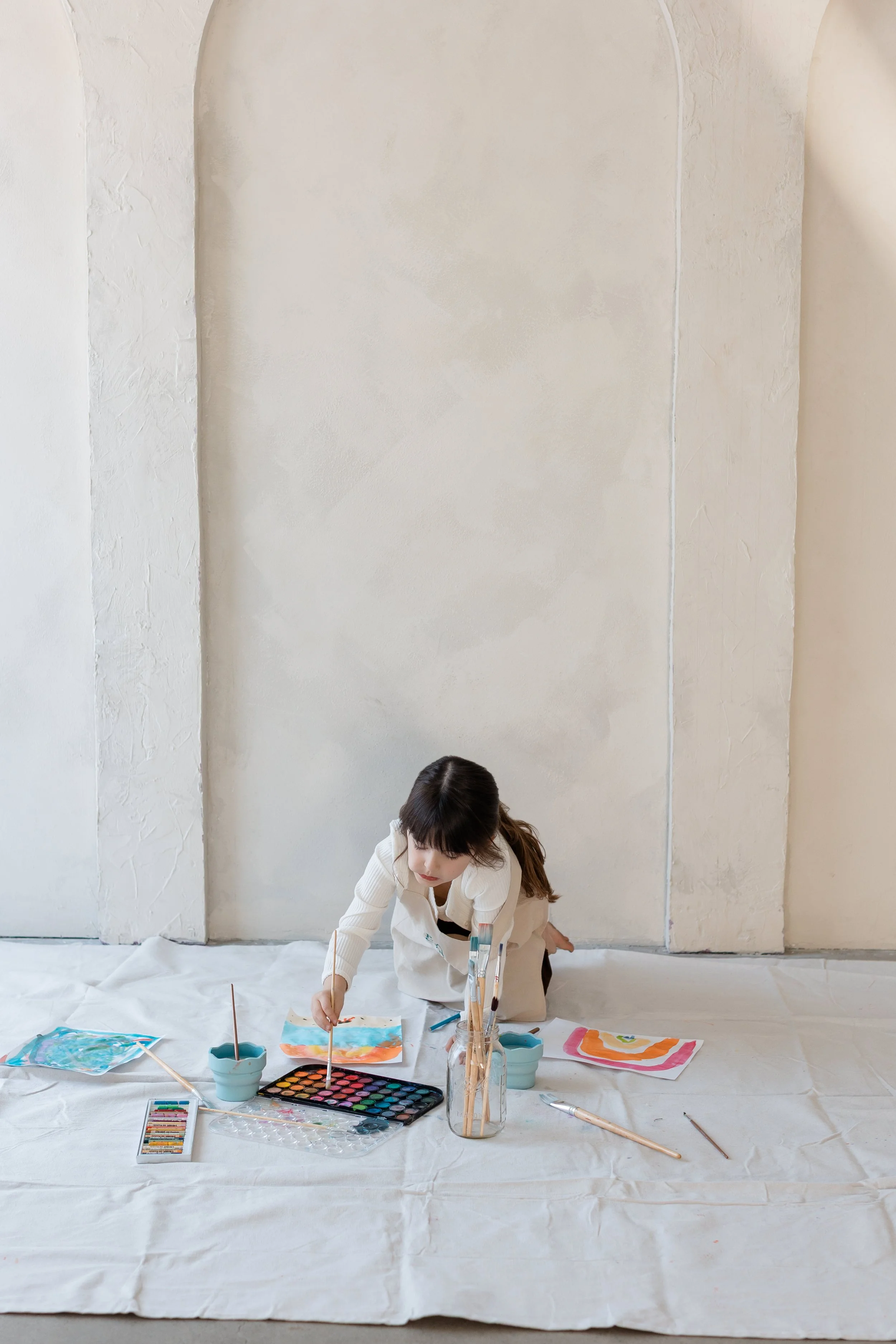 Young child painting with watercolors surrounded by brushes, paints, and finished artwork at Petite Canvas.