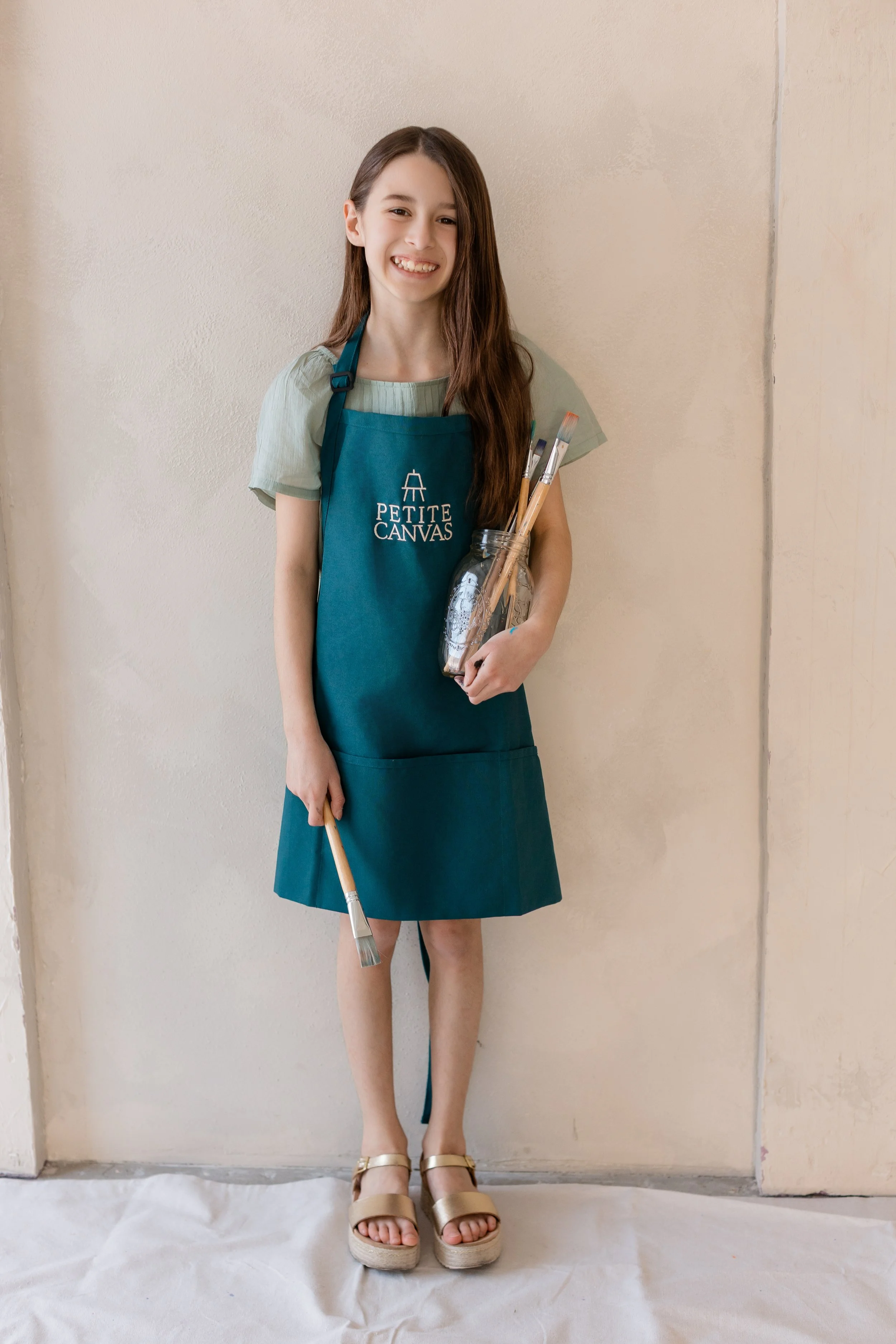 Young artist wearing a Petite Canvas apron and holding a jar of paintbrushes.