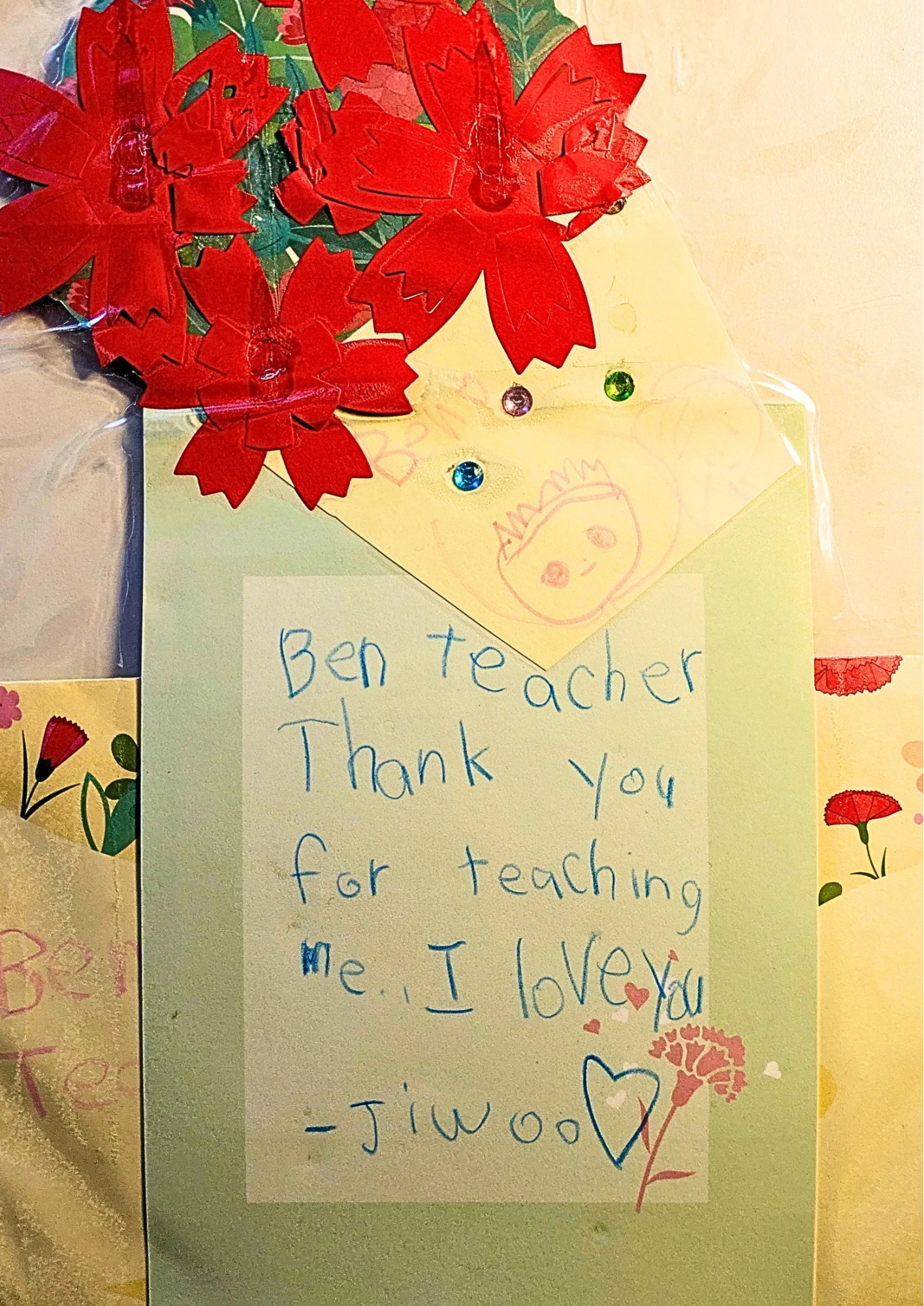 A handwritten note on yellow paper with colorful drawings and decorations, including red poinsettia flowers with gem accents, a smiling face, hearts, and floral designs. The note is a thank you message to a teacher from a student named Jiwon.