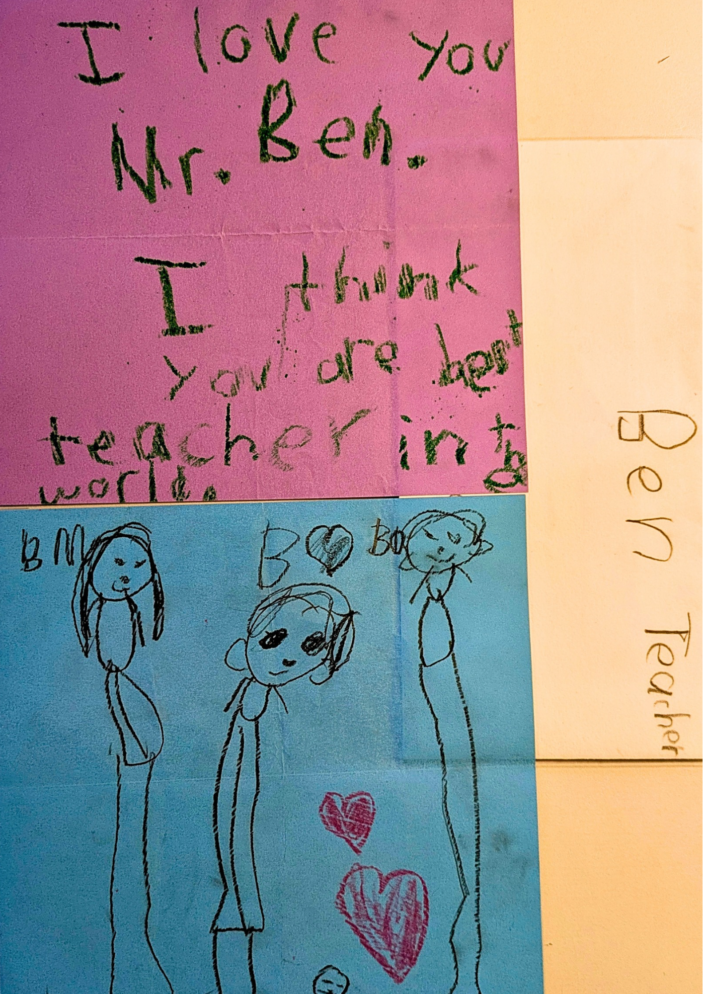 Colorful paper with handwritten note and drawings. Pink paper in the top half has a note in green crayon that says, "I love you Mr. Ben. Thank you teacher in the world." Blue paper below has four simple stick figure drawings of girls with hearts, one