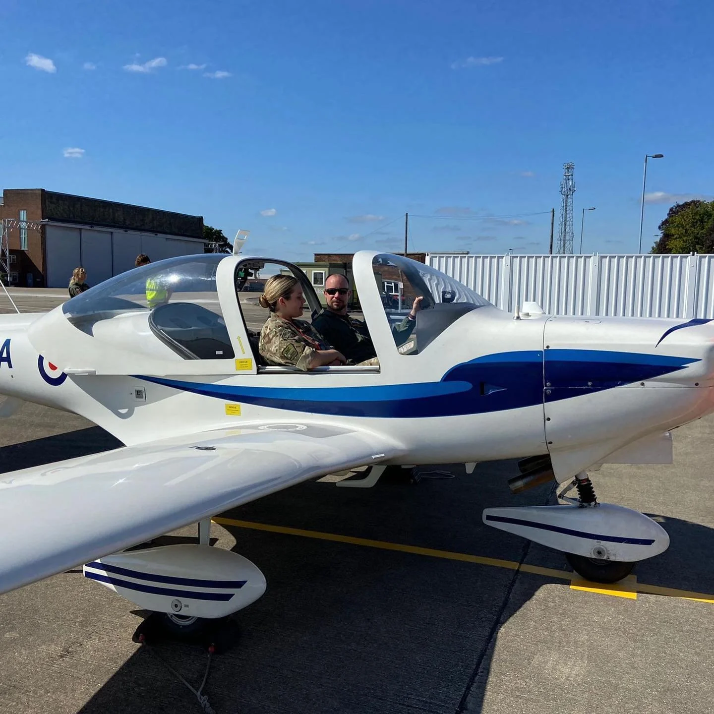 Two people sitting inside a small white and blue aircraft on a tarmac, with clear blue skies and some clouds in the background.
